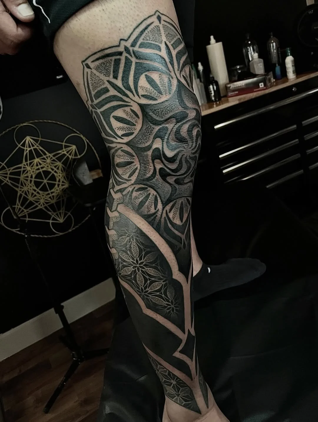 A person's arm with a large, intricate black tattoo featuring geometric and floral patterns extending from the shoulder to the wrist.