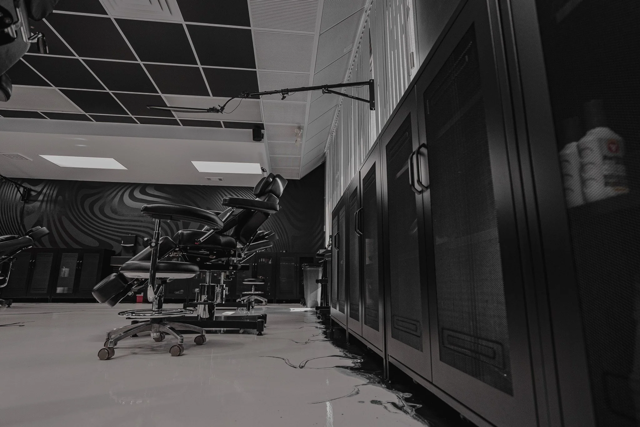 A modern indoor tattoo studio with black tattoo chairs, black storage cabinets, and a dark geometric wall design.
