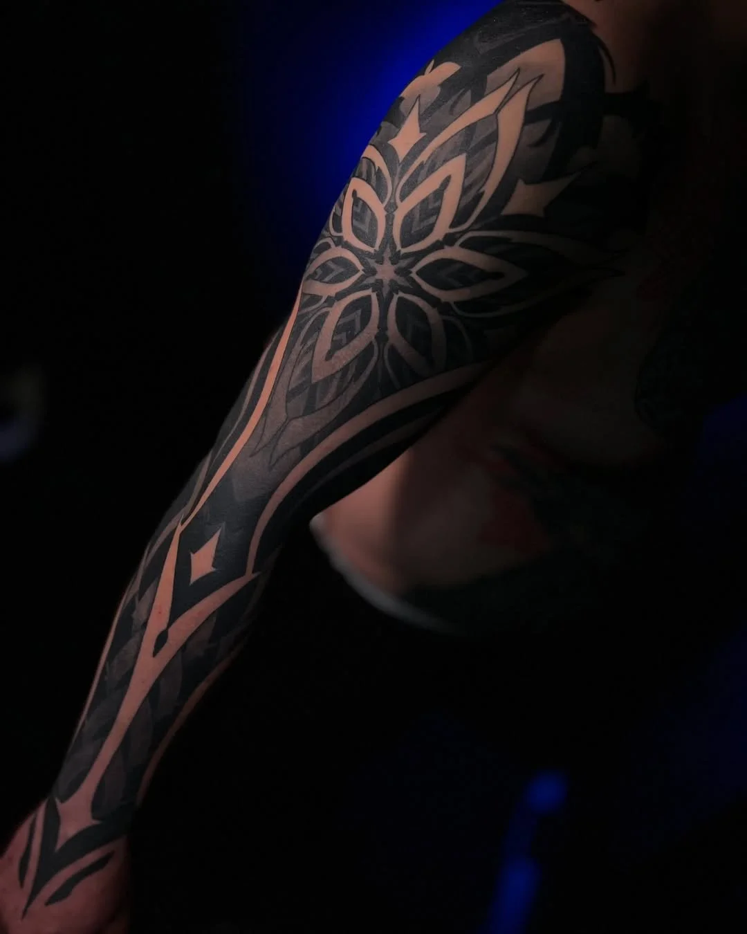A person's arm with an intricate black tattoo featuring a geometric floral pattern, set against a dark background with faint blue lighting.