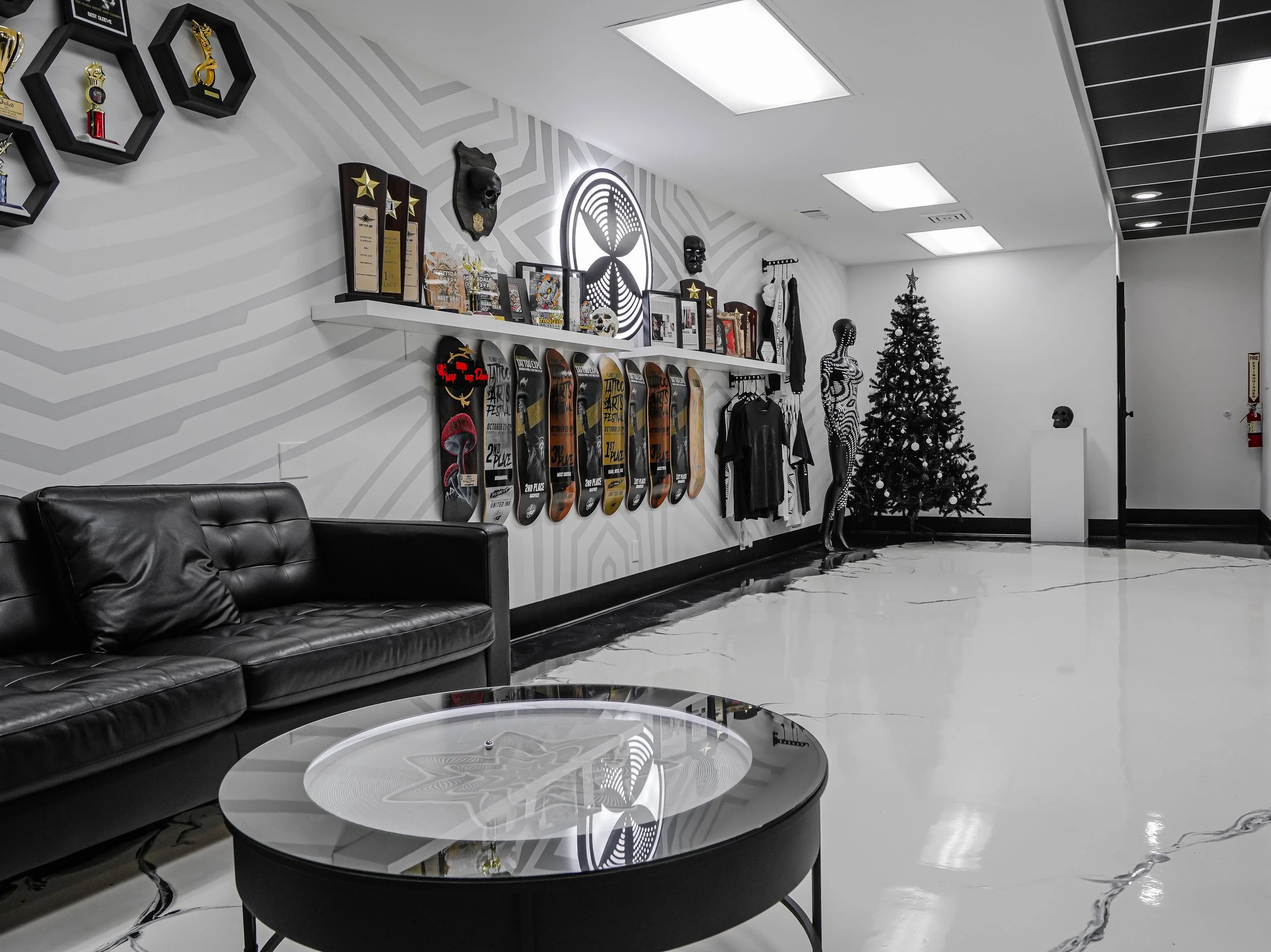 Modern lobby with black leather sofa, glass coffee table, white shelves with trophies and skateboard decks, mannequins, a decorated Christmas tree, and art pieces on the wall.