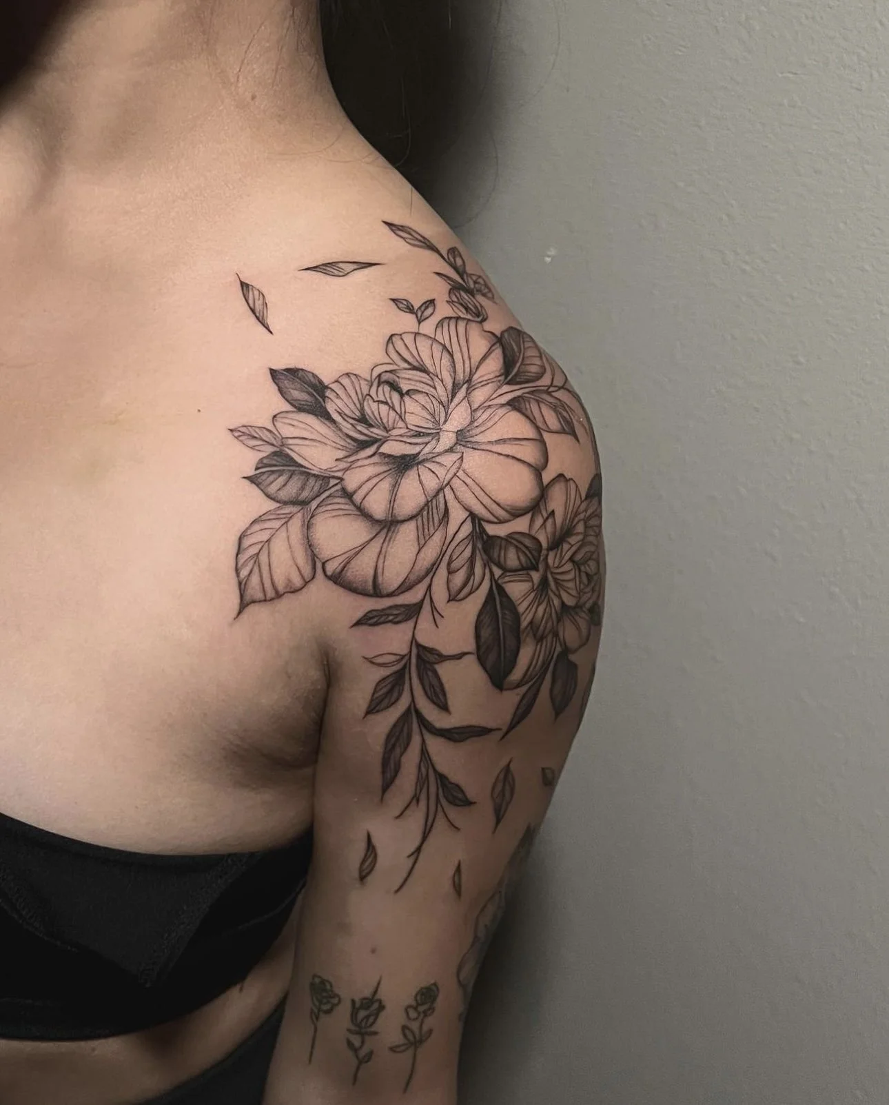 A detailed black and gray floral tattoo on a person's shoulder and upper arm, featuring large blooming flowers, leaves, and small buds.