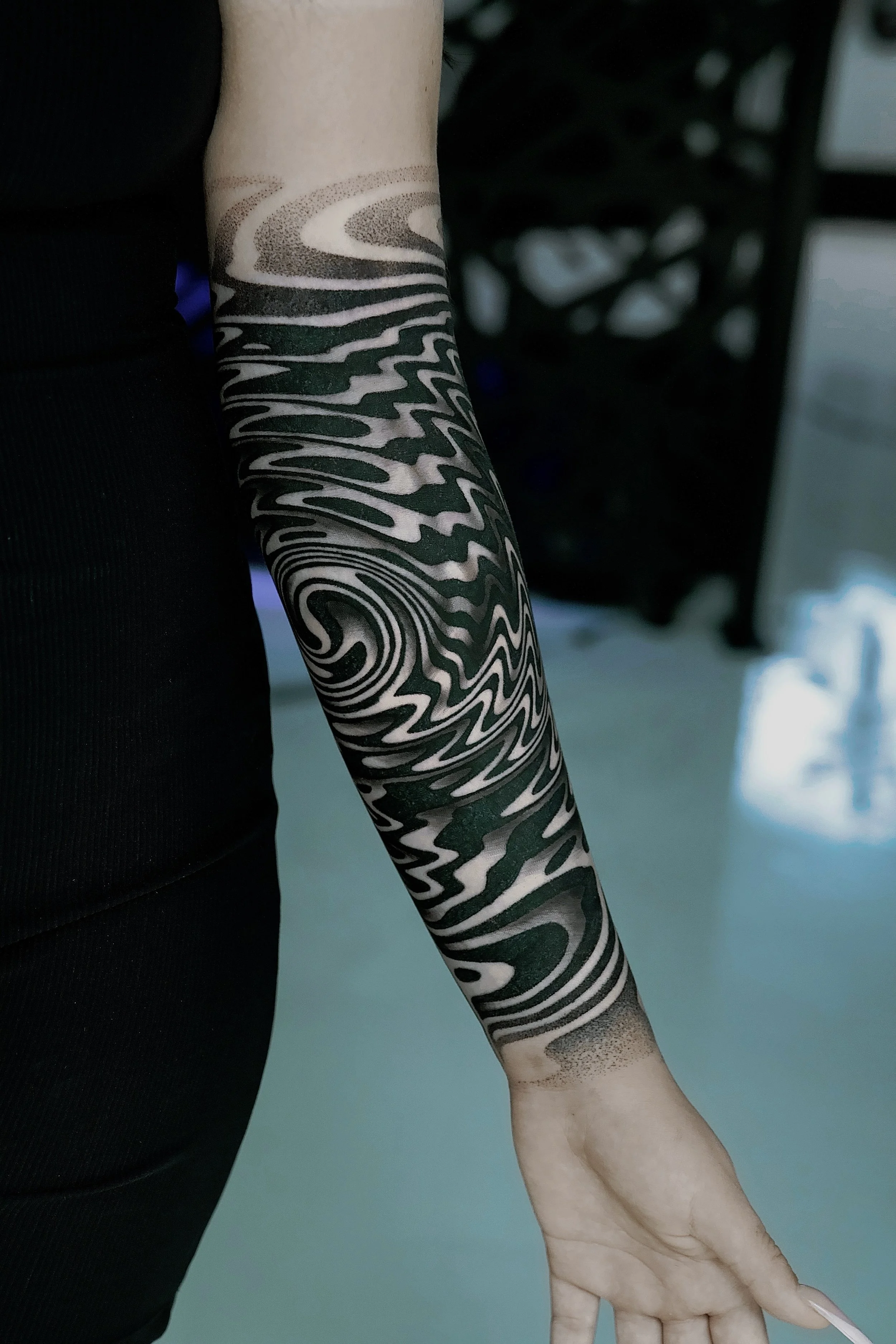 A person's arm with a black and beige swirling tattoo extending from the wrist to the elbow, featuring intricate, abstract patterns.