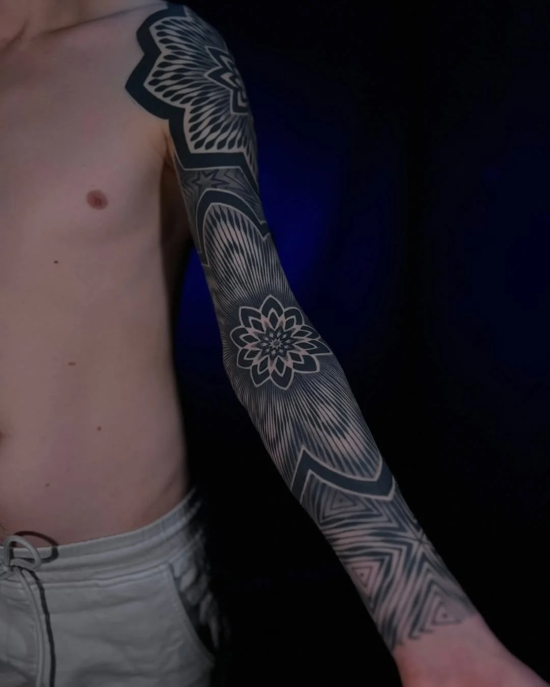 A person's left arm with a detailed black and white tattoo sleeve featuring geometric and floral patterns, extending from the shoulder to the wrist. The person is shirtless, wearing light-colored shorts, against a dark background.