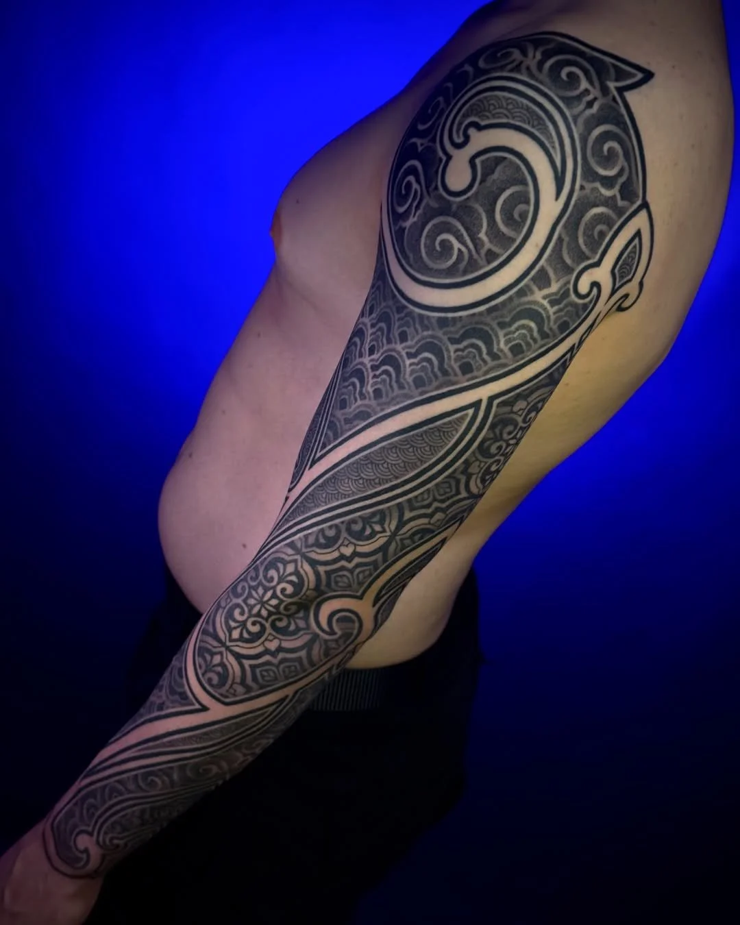Person with Polynesian-style black tattoo sleeve on their arm, against a dark blue background.
