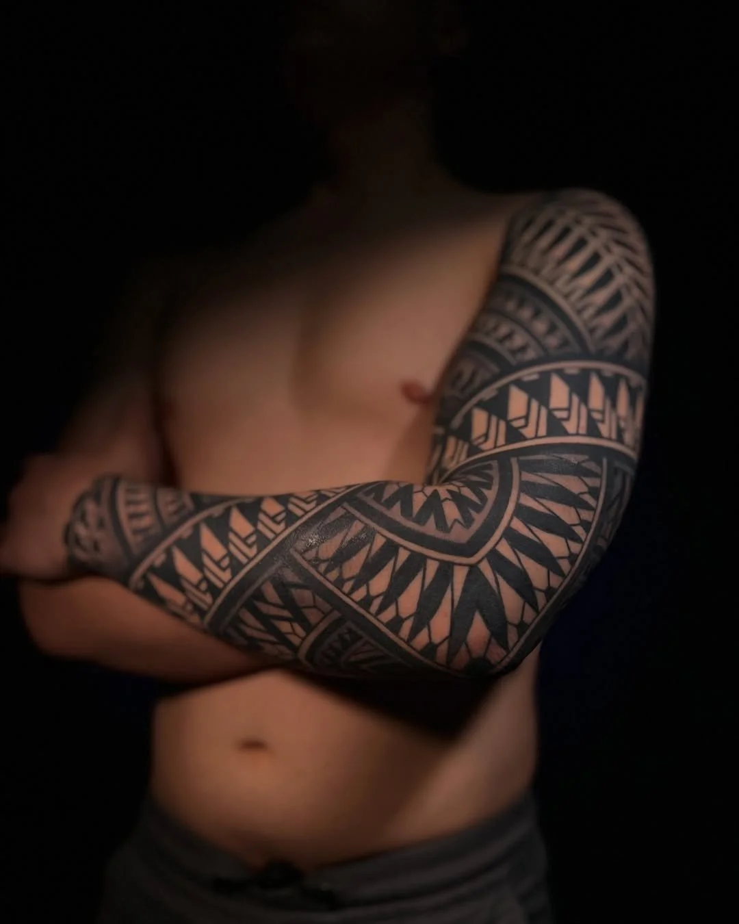 A person with a large intricate black and grey tattoo sleeve on their arm, bare-chested against a dark background.