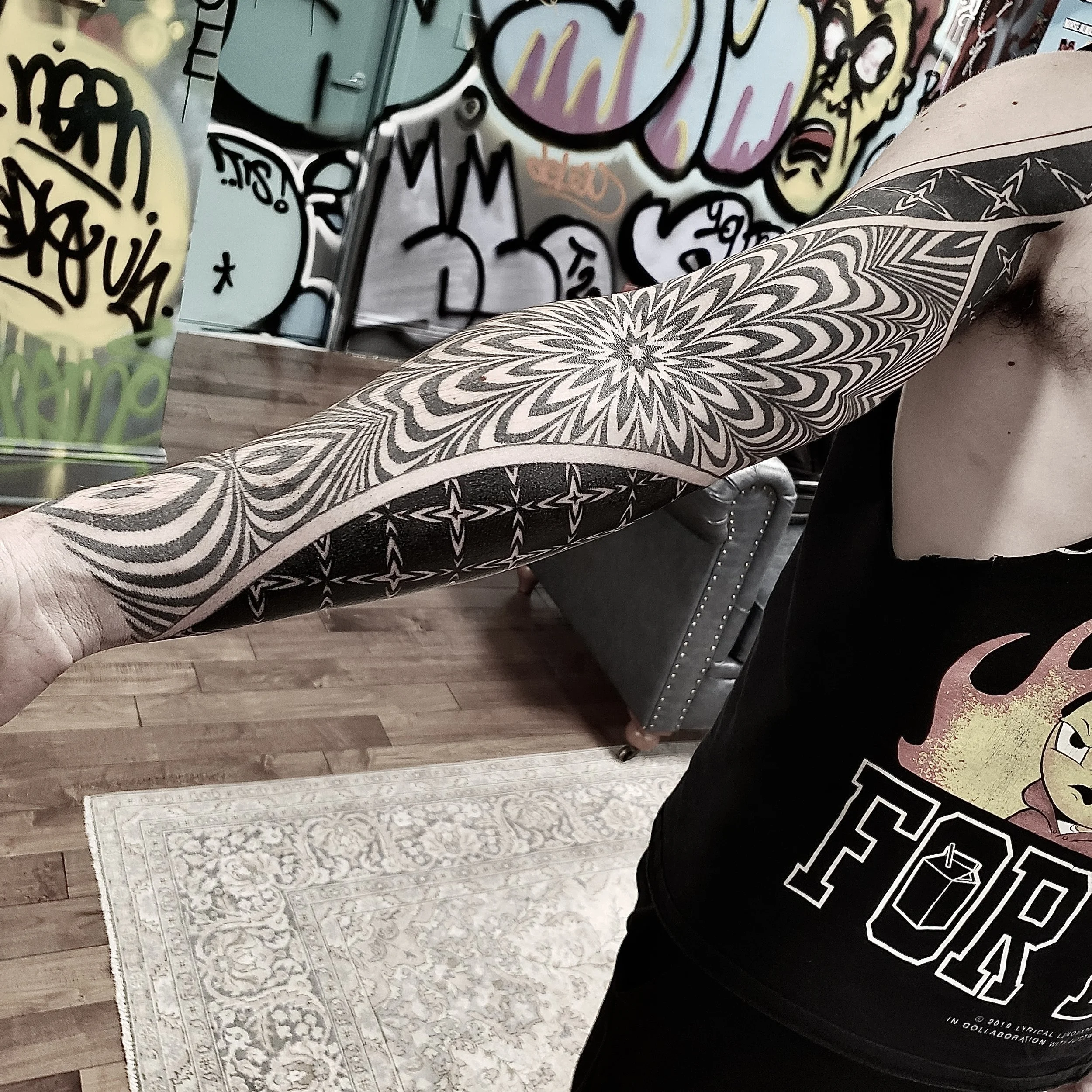 A person with a tattooed arm extending towards the camera, showing an intricate black and gray geometric tattoo. The background features colorful graffiti art on a wall, a gray upholstered couch with nailhead trim, and a patterned area rug on wooden 