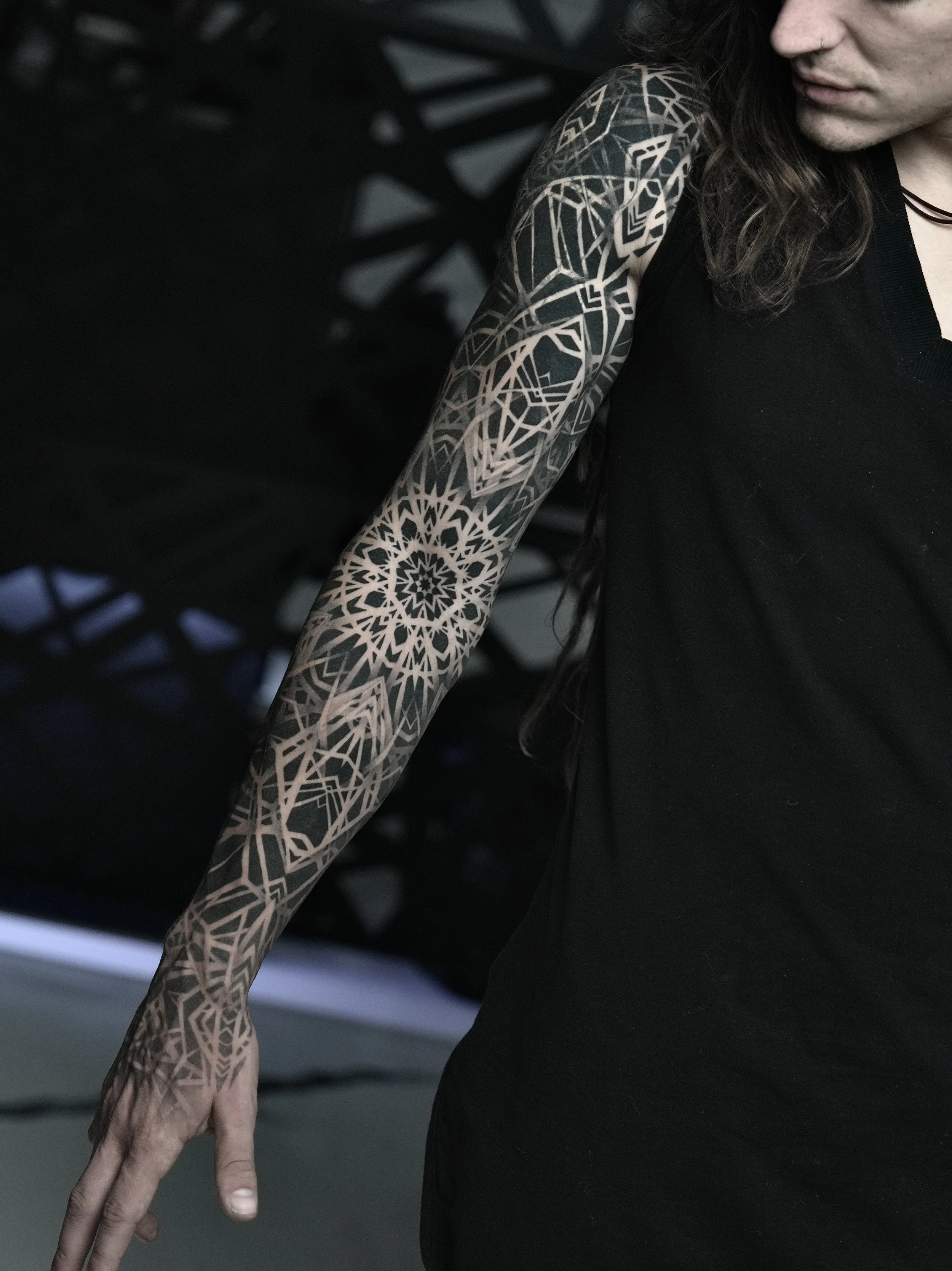 A person with long dark hair and light skin wearing a black sleeveless shirt, displaying a detailed black and beige tattoo sleeve on their left arm, featuring geometric patterns and floral motifs.