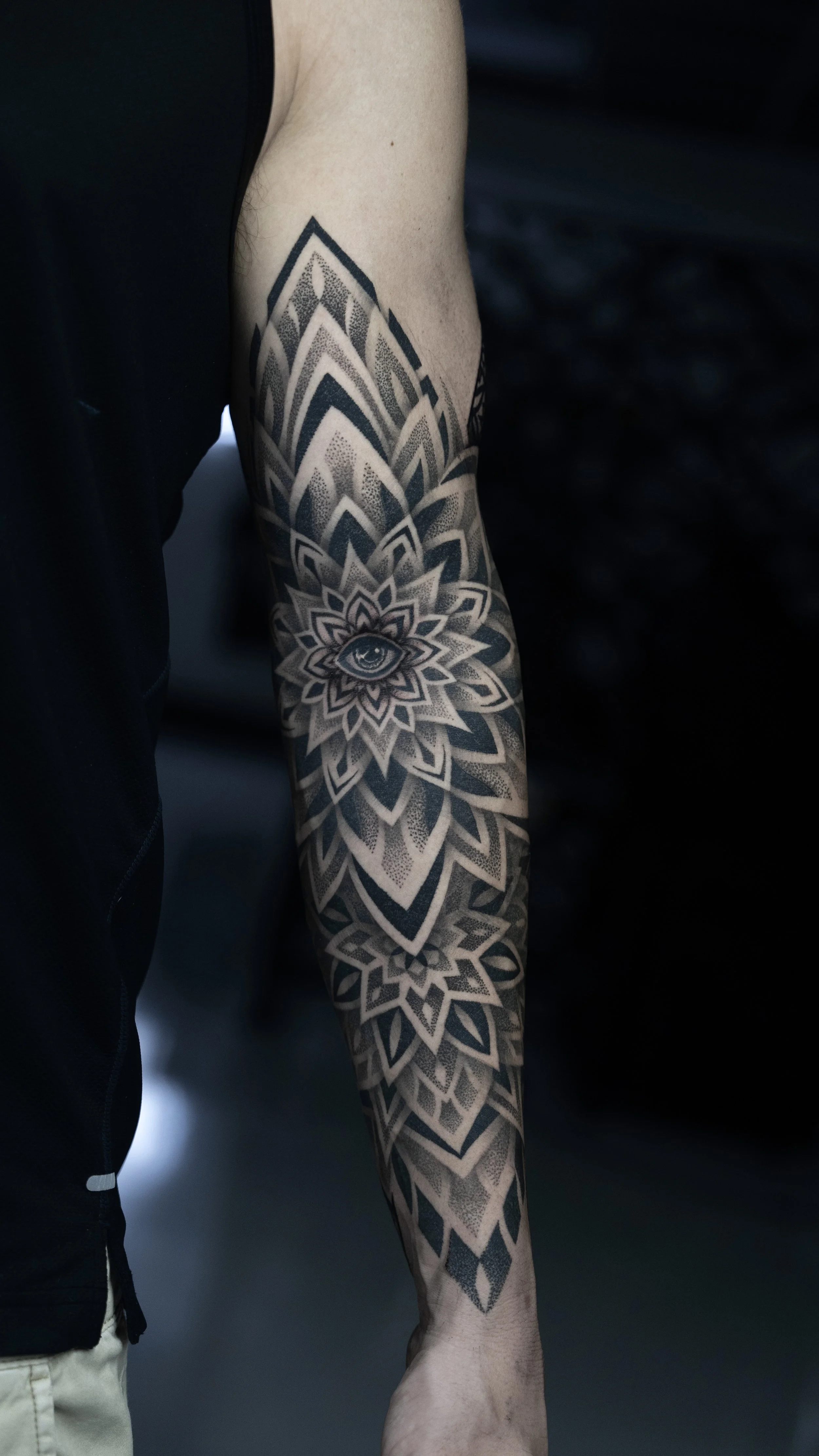 A person's arm with a detailed black and gray tattoo sleeve featuring geometric patterns and an eye within a floral design.