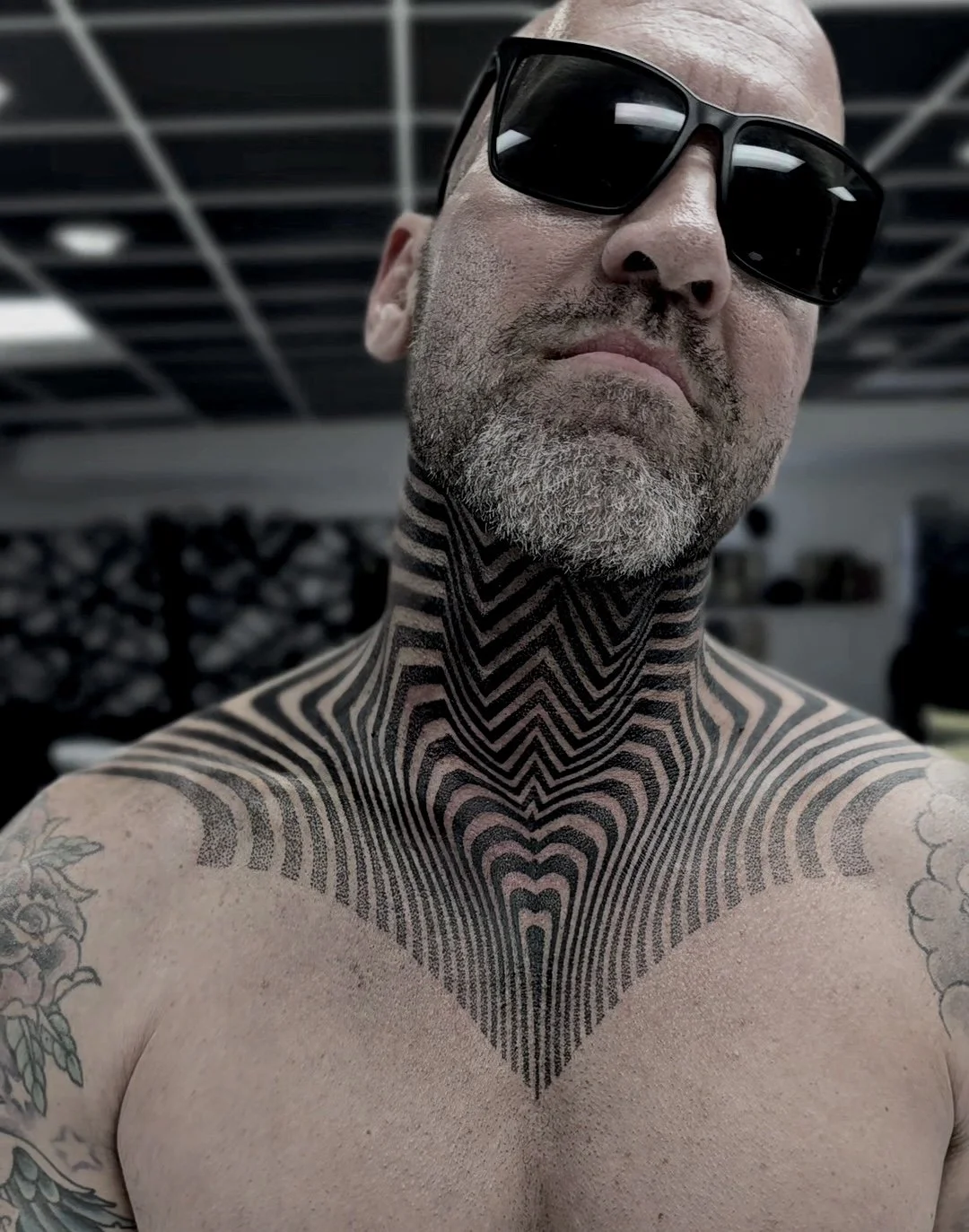 A bald man wearing large black sunglasses, with a gray beard, showing a geometric black tattoo on his neck and upper chest, and floral tattoos on his shoulders.