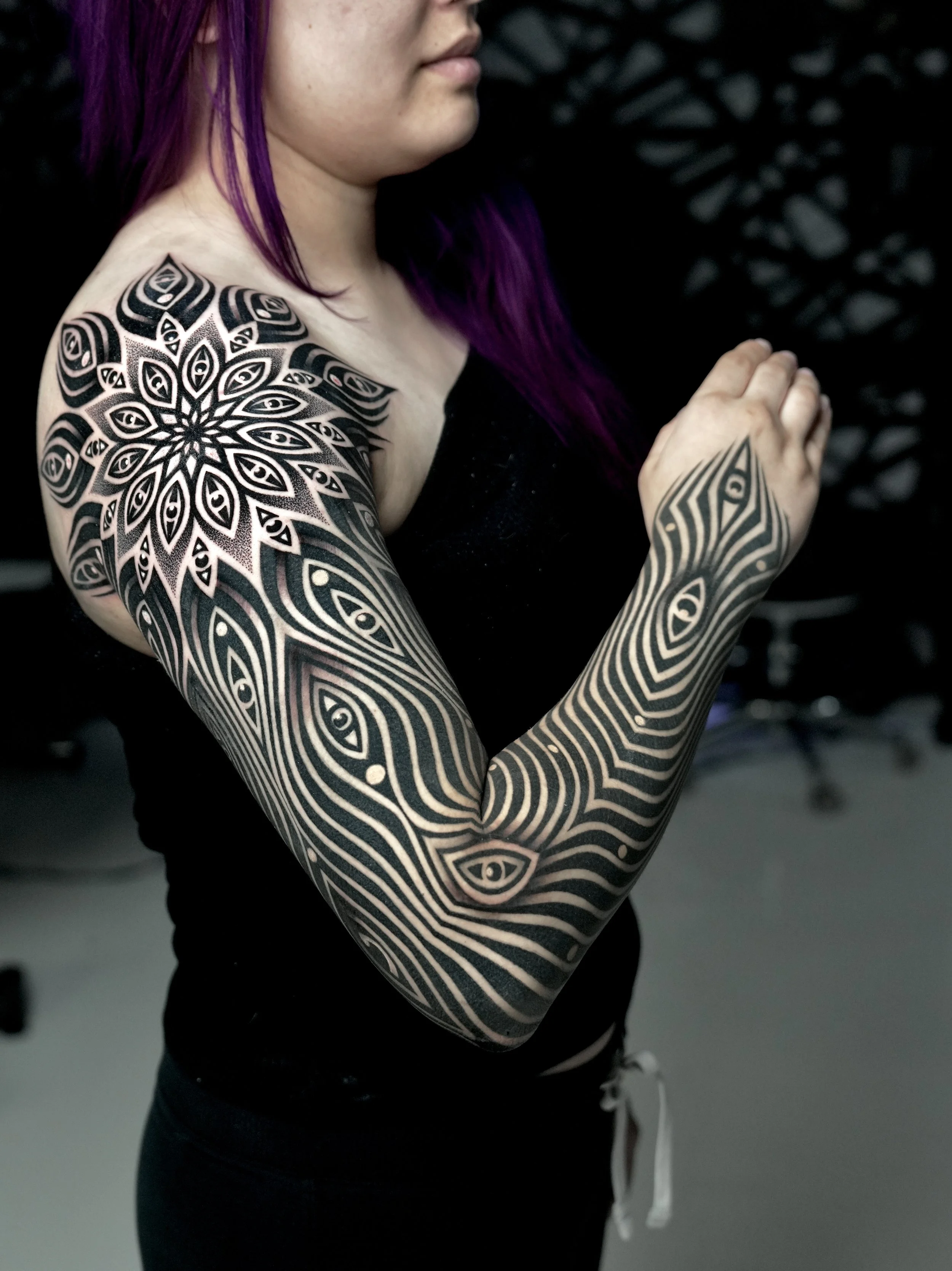 A woman with purple hair and elaborate black and white geometric tattoos on her arm and shoulder, standing indoors against a dark background.