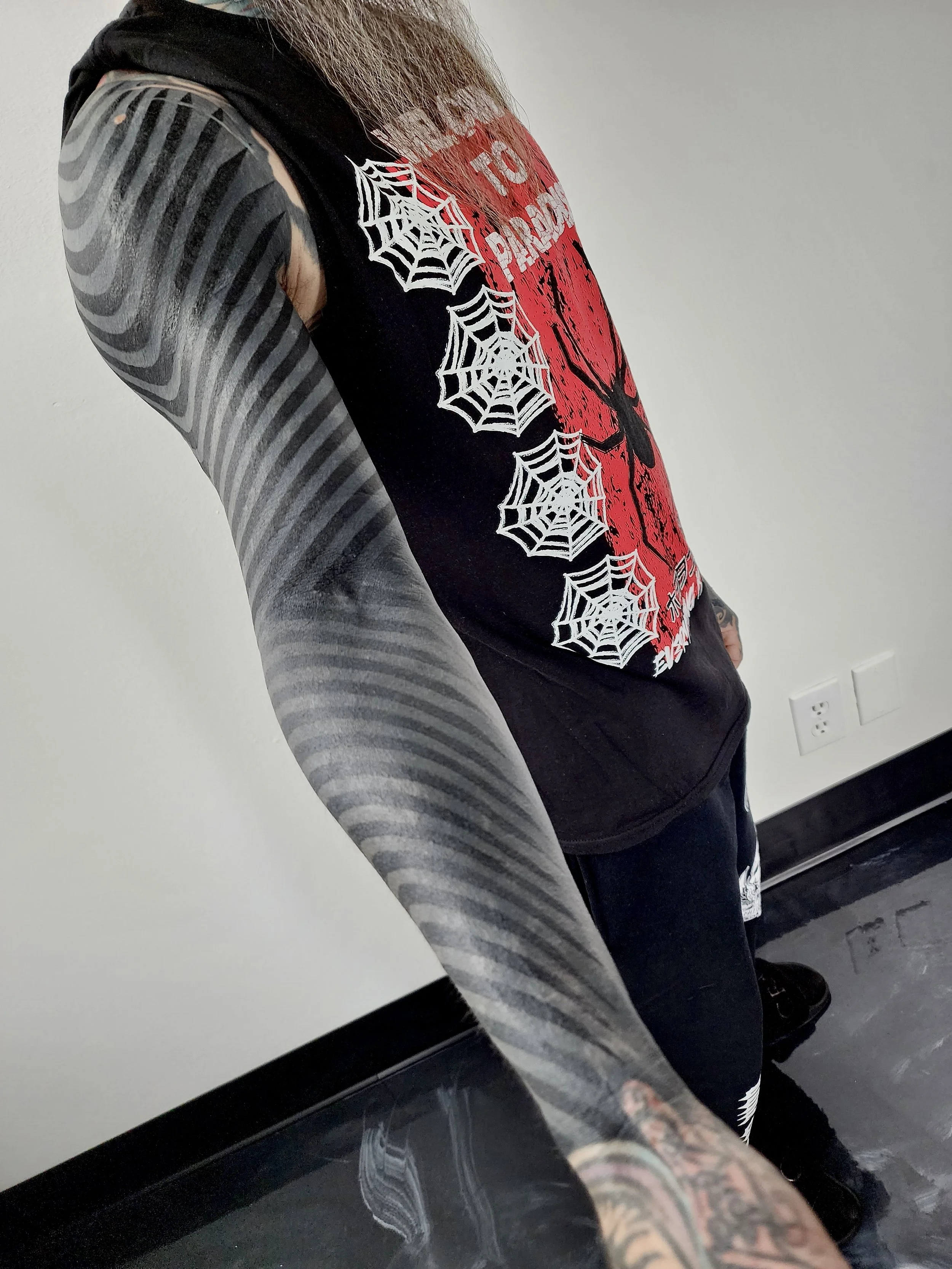 Person wearing a black sleeveless shirt with white spider web designs and a red graphic, displaying striped arm tattoos and tattoos on the hand.