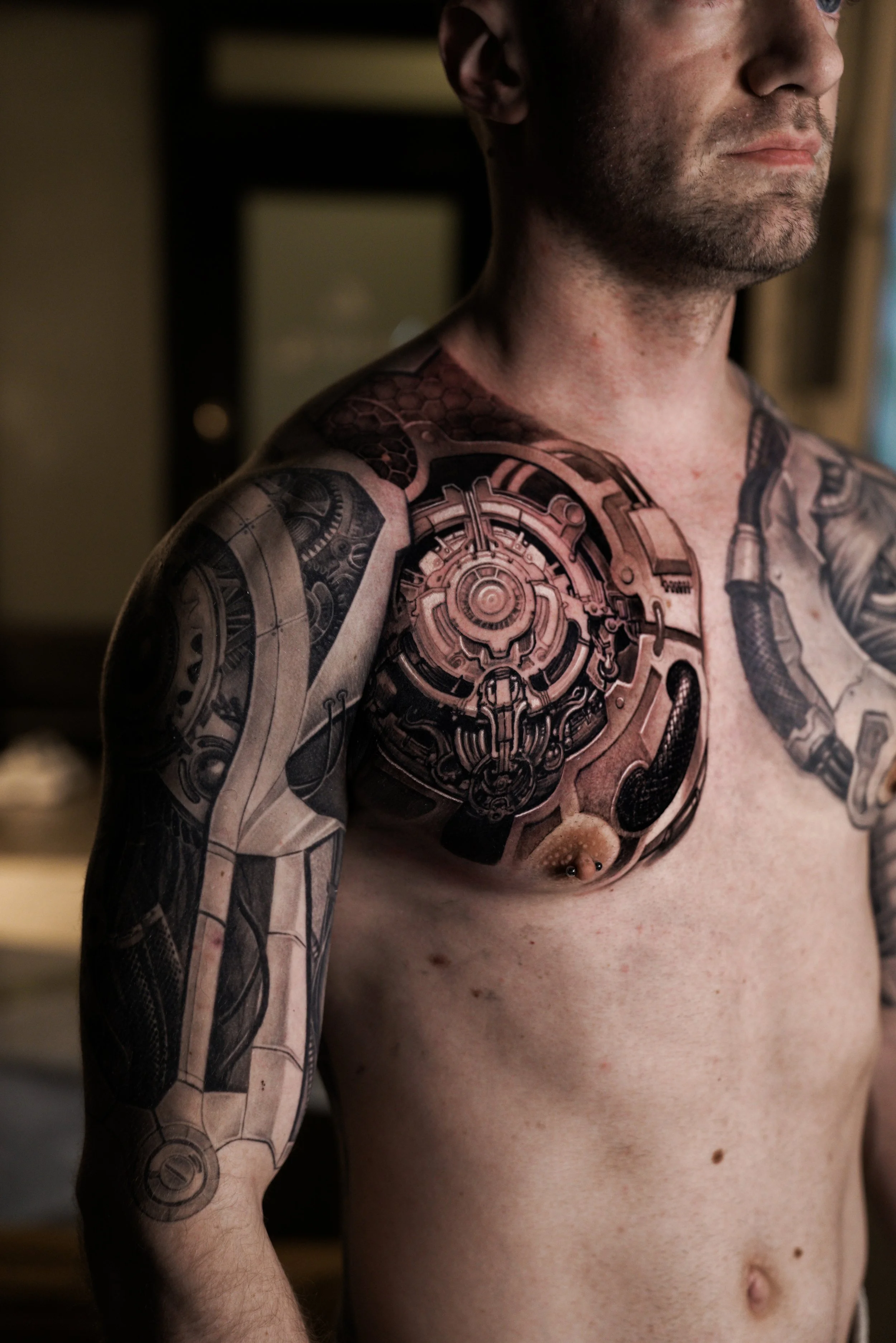 A man with tattoos covering his shoulder, chest, and arm, showing mechanical and futuristic designs, standing shirtless in an indoor setting.