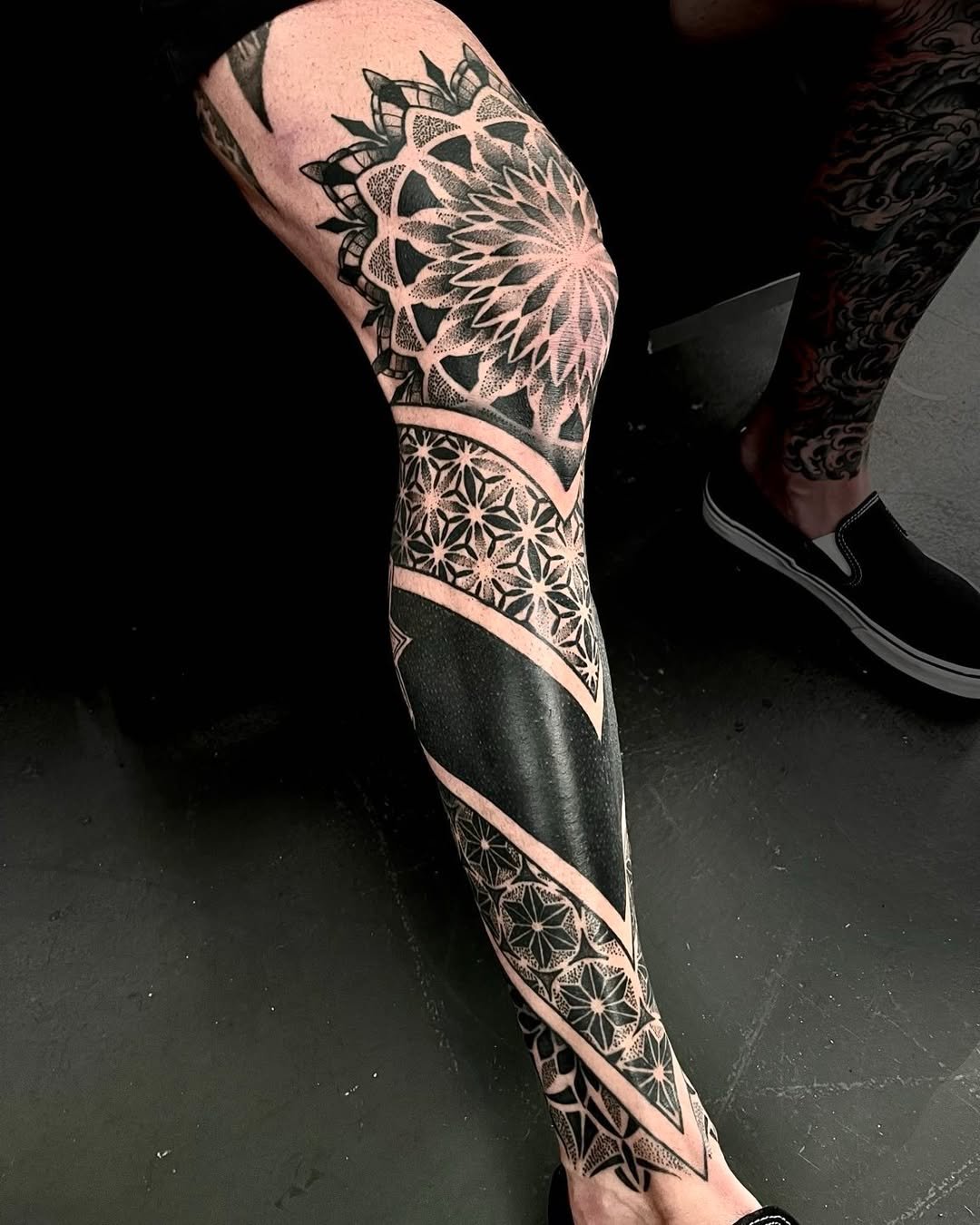 A person's leg with a large geometric black and white tattoo covering the thigh, knee, and lower leg, featuring intricate patterns and symmetrical designs.