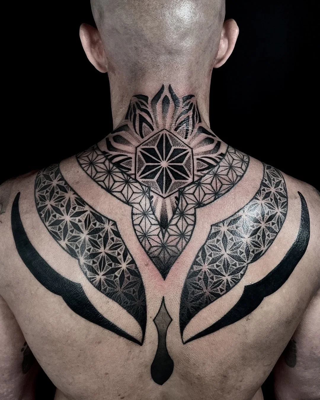 A person's back and neck with a large, black geometric tattoo extending from the neck to the chest, featuring intricate patterns and symmetrical designs.