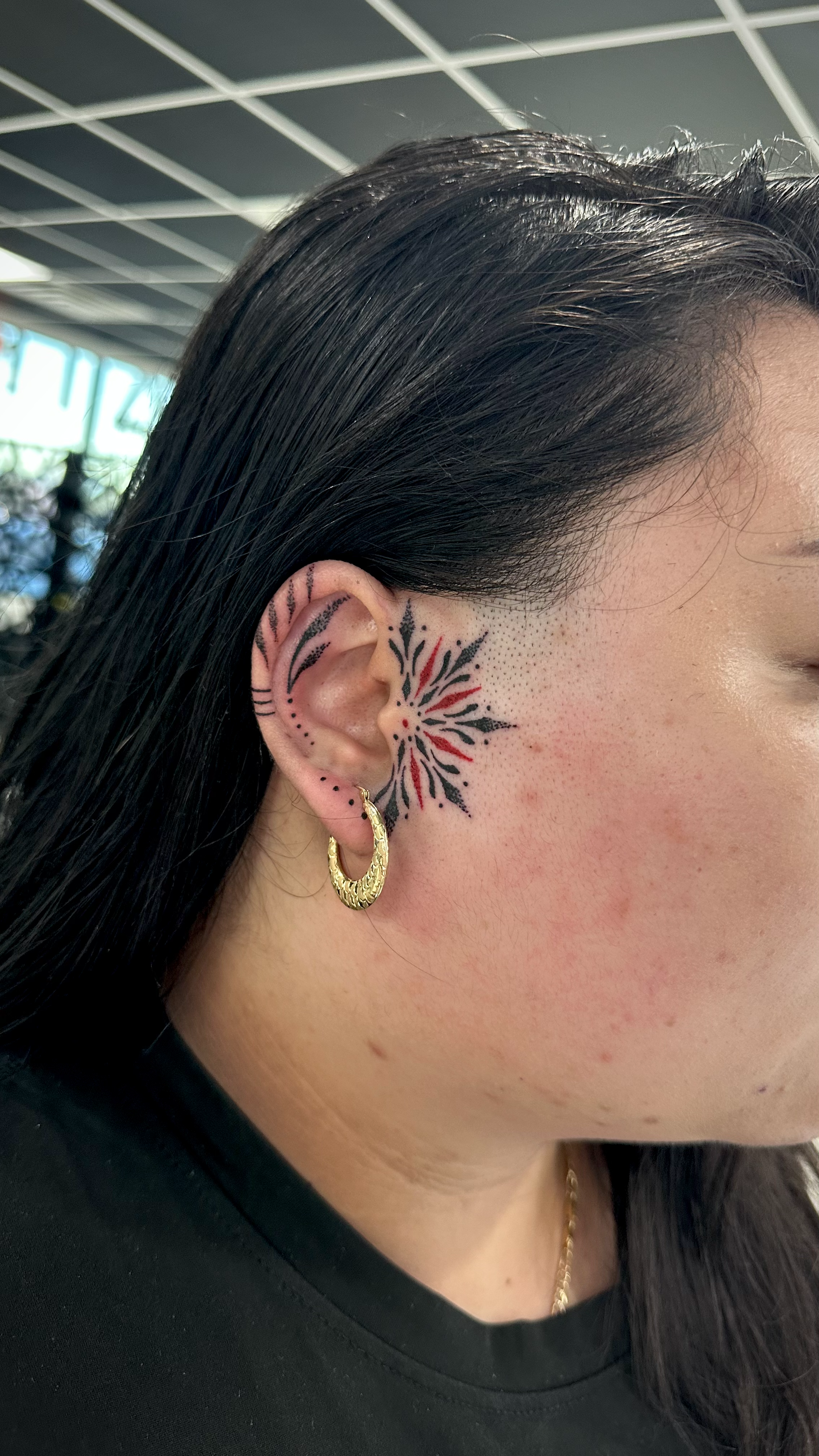 Close-up of a woman's face showing tattoo behind her ear and gold hoop earring, with straight black hair and a black shirt.