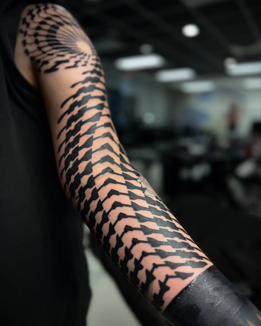 Close-up of a person's arm with a black tribal tattoo covering the entire arm, in an indoor setting.