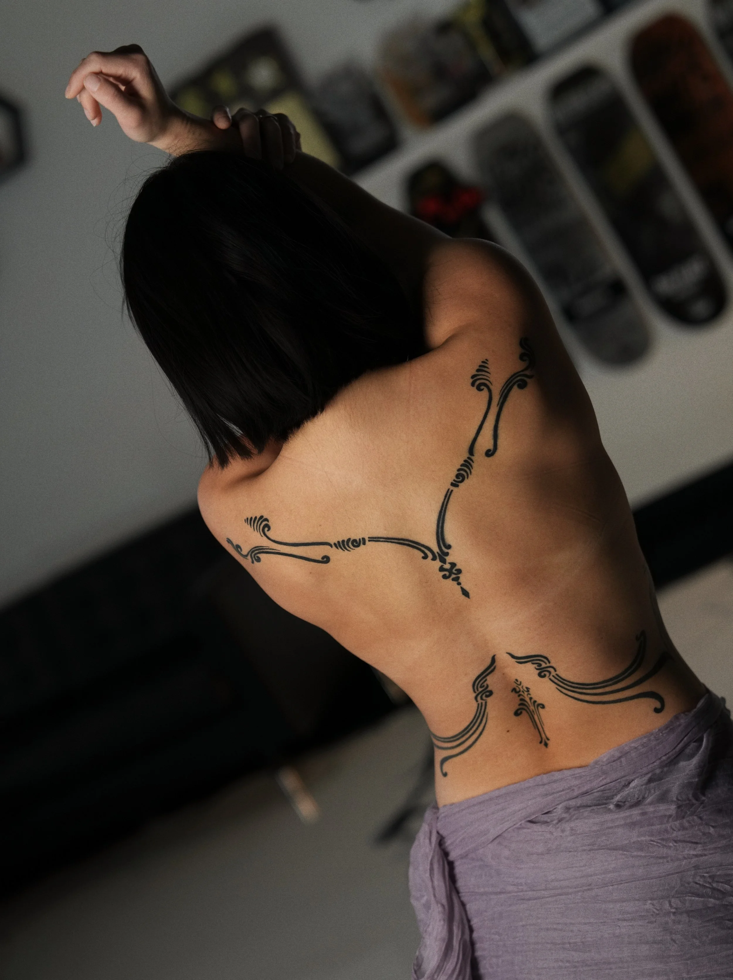 A woman with short, dark hair showing her back and shoulder tattoos, wearing a purple skirt, posed indoors with her arm raised.