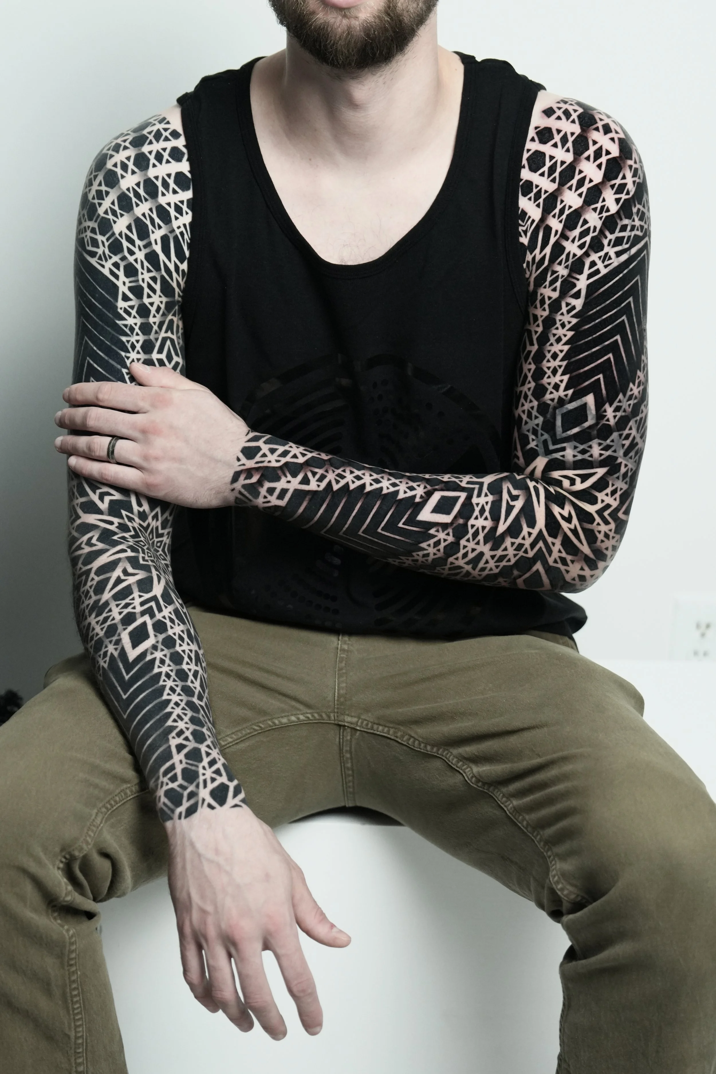 A person sitting with arms crossed, wearing a black sleeveless shirt with tattoos on both arms, and beige pants.