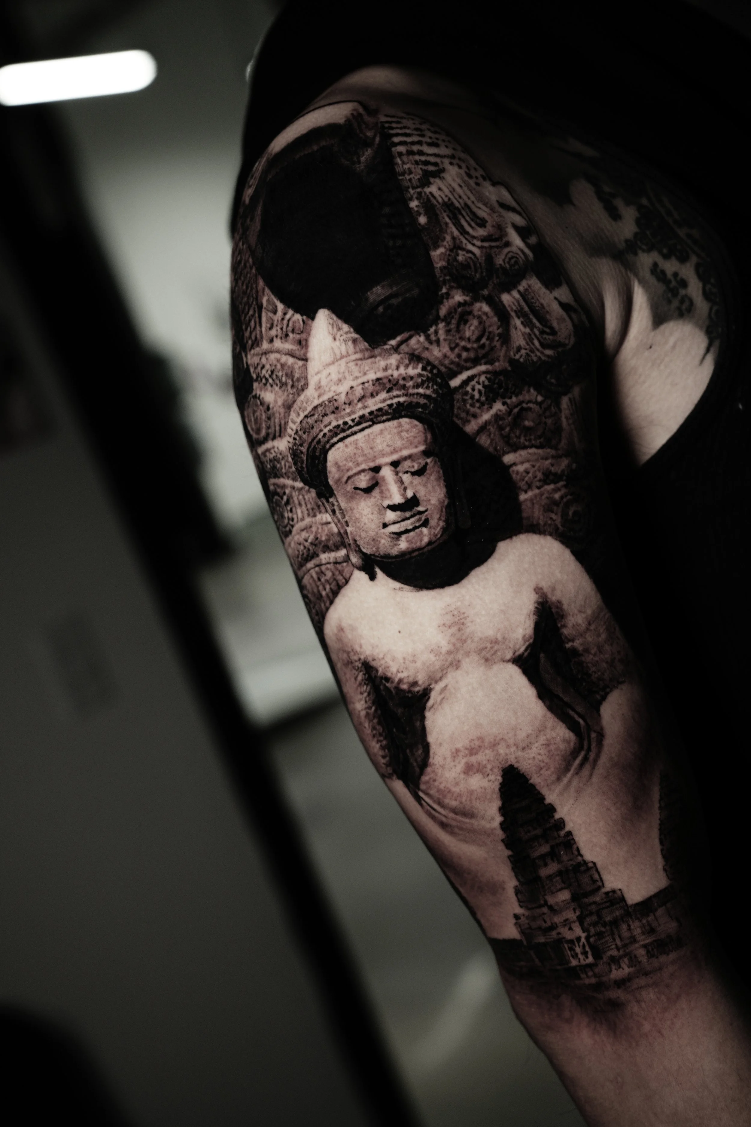 Close-up of a person with a large tattoo on their upper arm, featuring a woman's face with closed eyes and decorative elements, in black and gray ink.