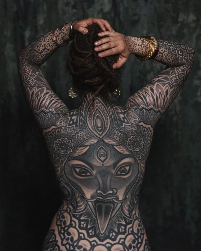 Back of a person with intricate black tattoos covering their back, arms, and shoulders, featuring a symmetrical design with a central mask or face, floral patterns, and geometric shapes. The person is holding their head with both hands, and they are 