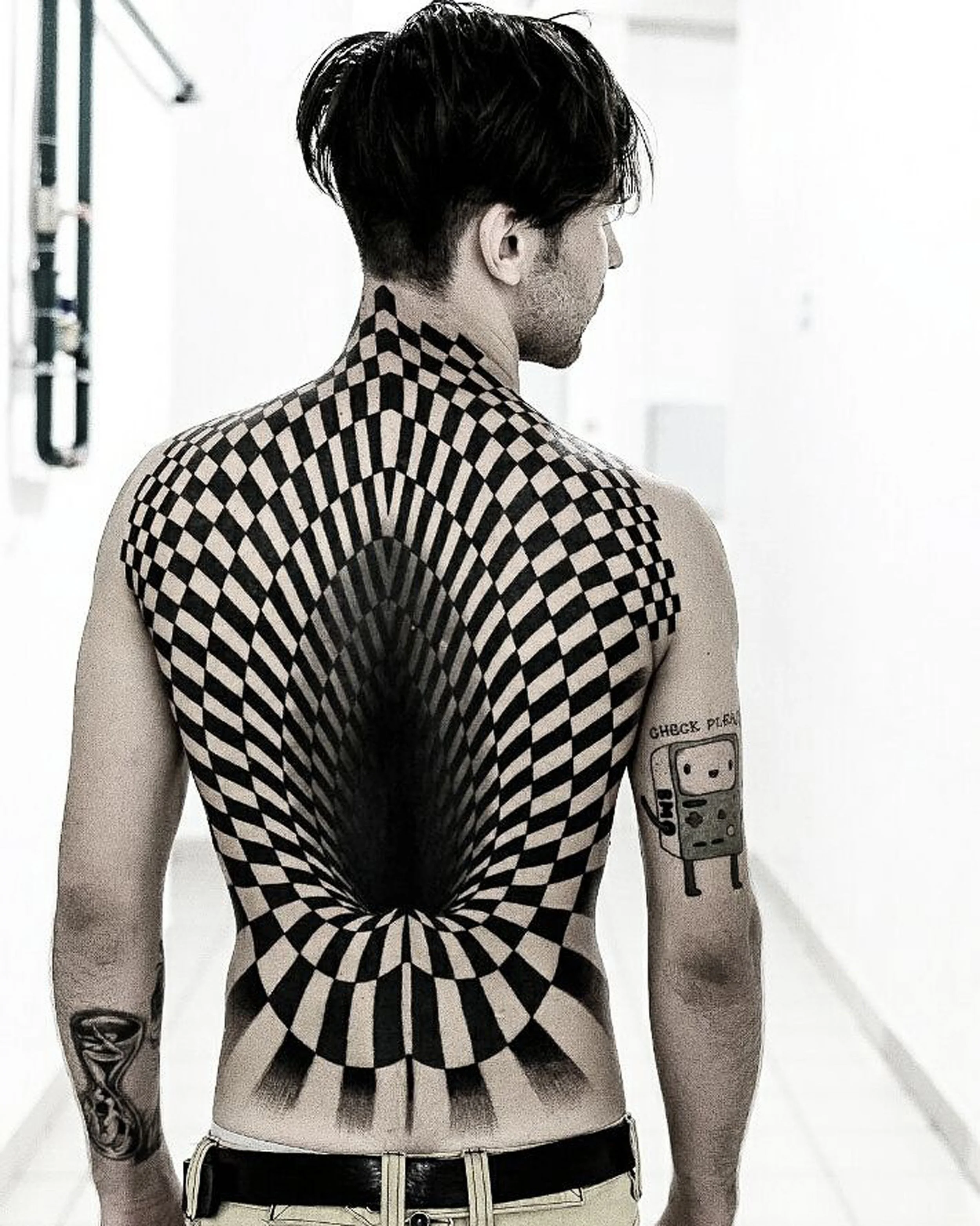 A man with black hair and multiple tattoos on his left arm, looking to the right, in a room with white walls.