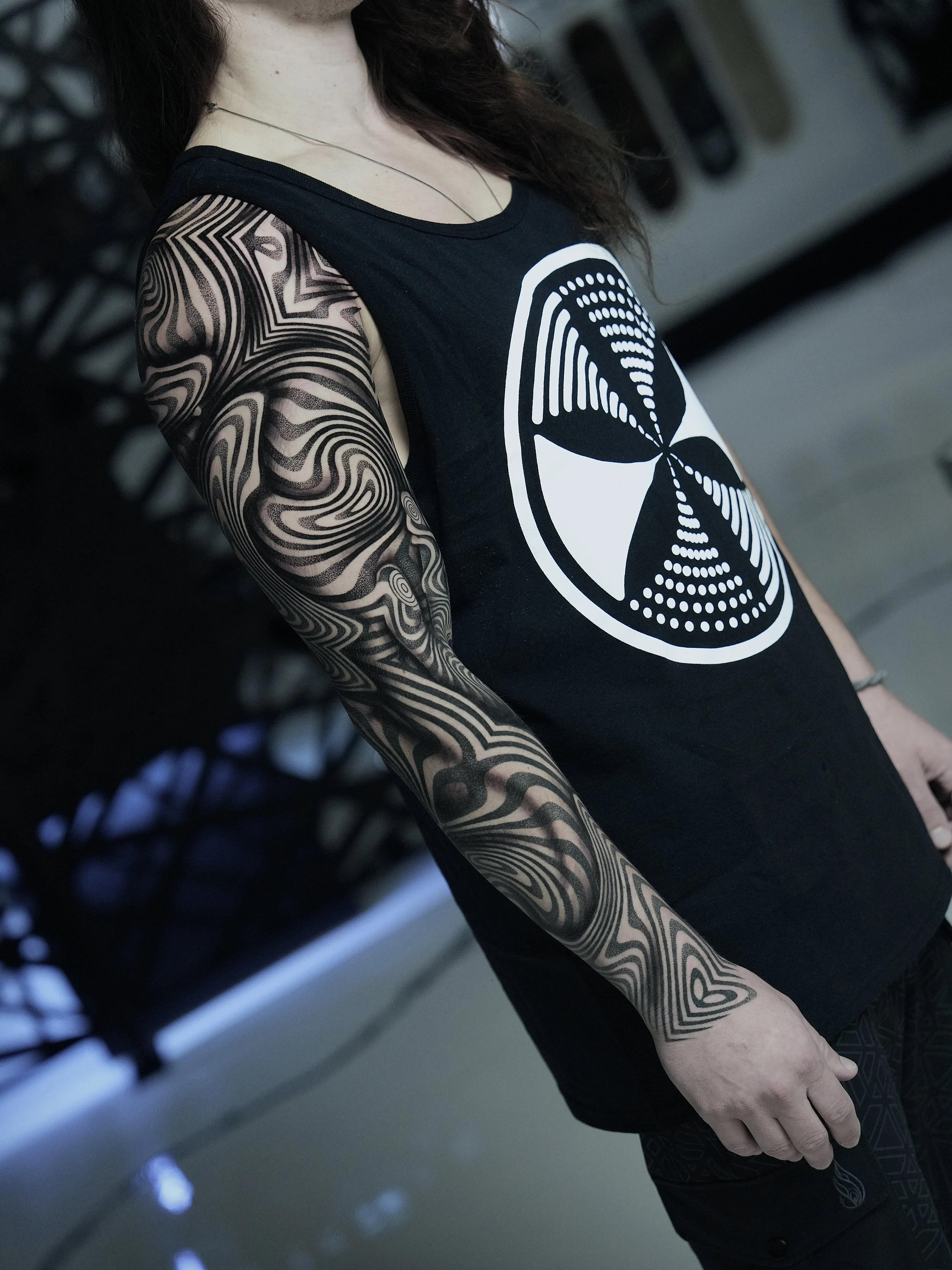 Person with long dark hair wearing a sleeveless black shirt with a white geometric logo and intricate black and gray sleeve tattoo.