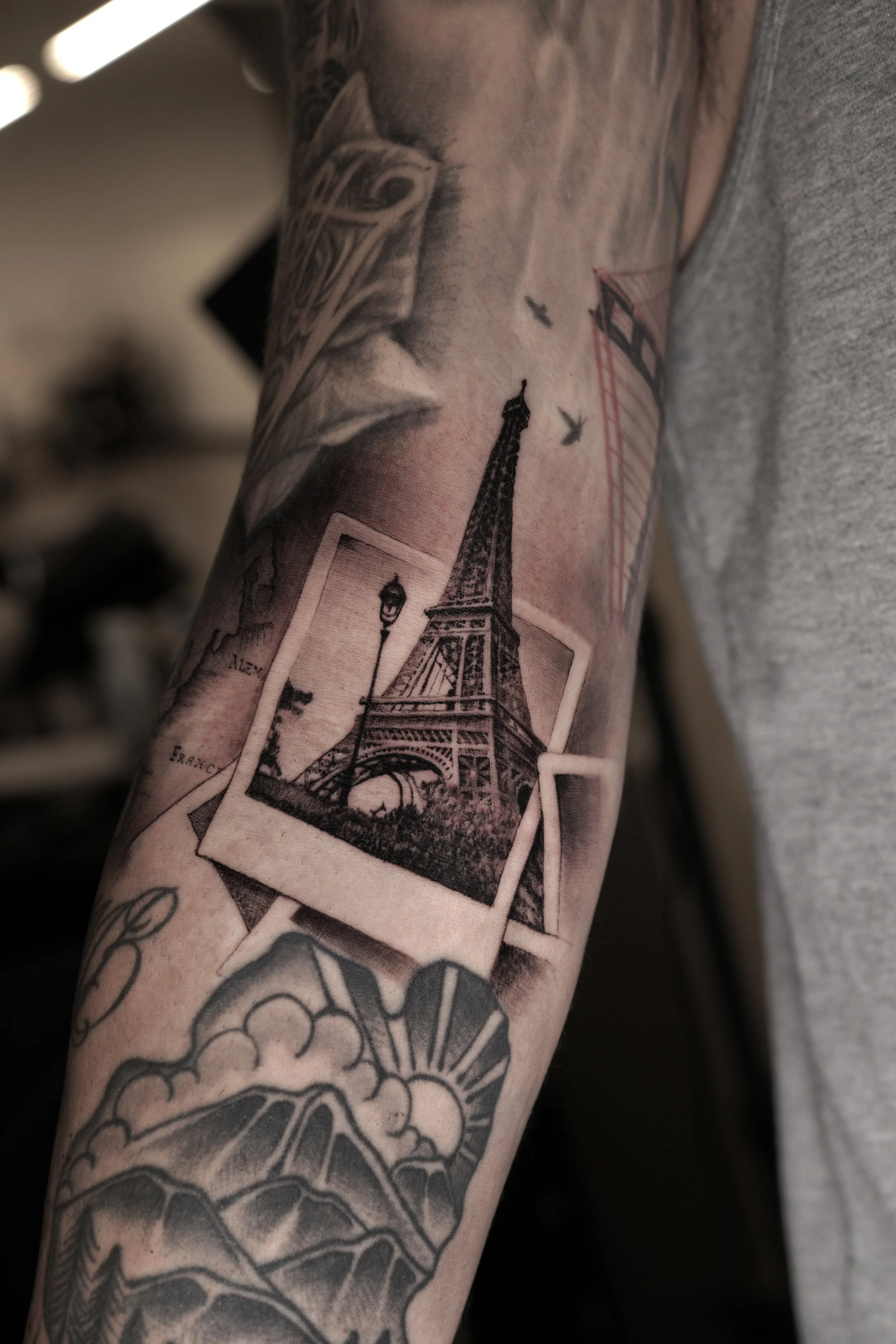 Black and gray tattoo of the Eiffel Tower, birds flying, and Parisian street lamps on a person's arm.