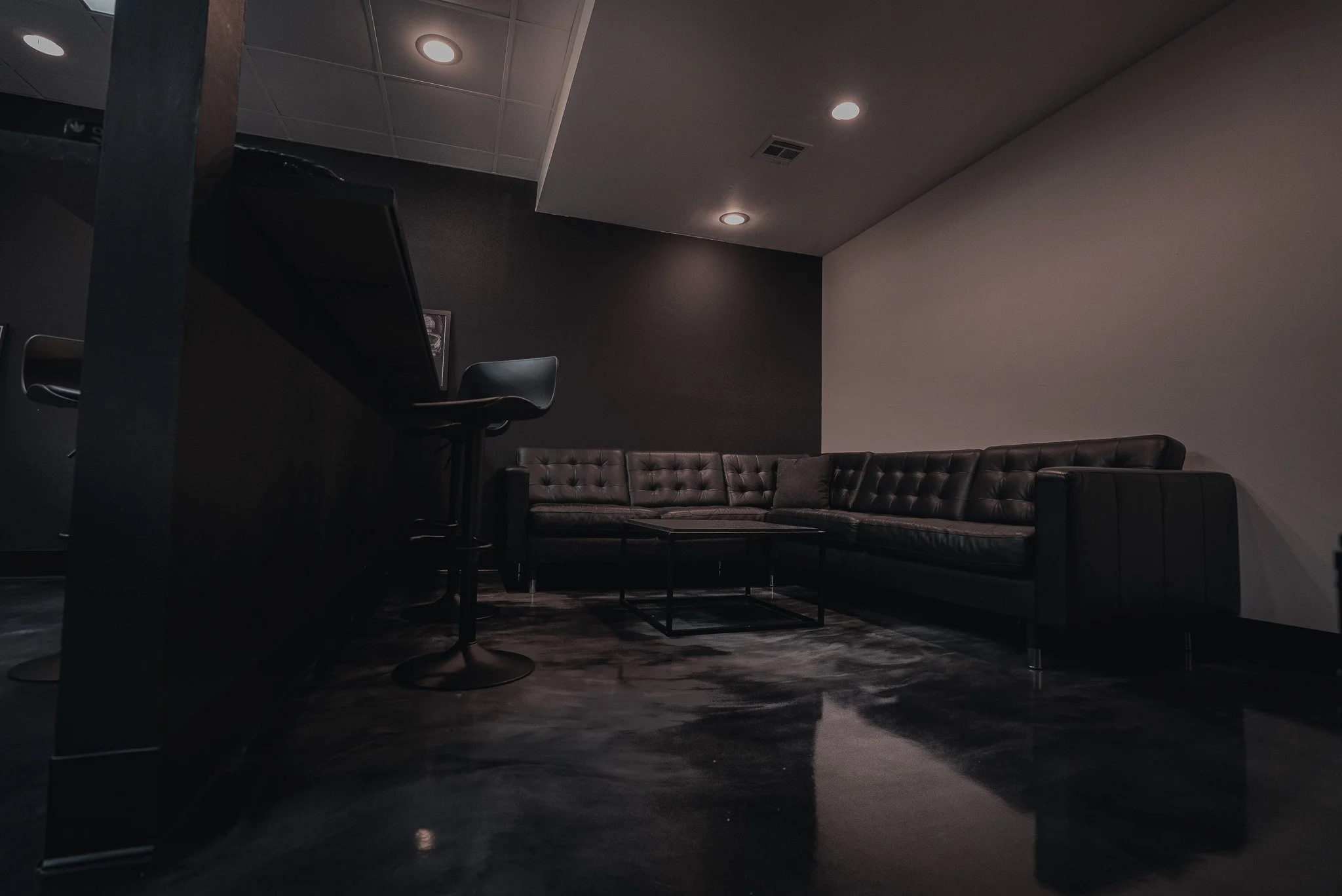 A dimly lit room with a black corner sofa, a small central table, a bar counter with barstools, and recessed ceiling lights.