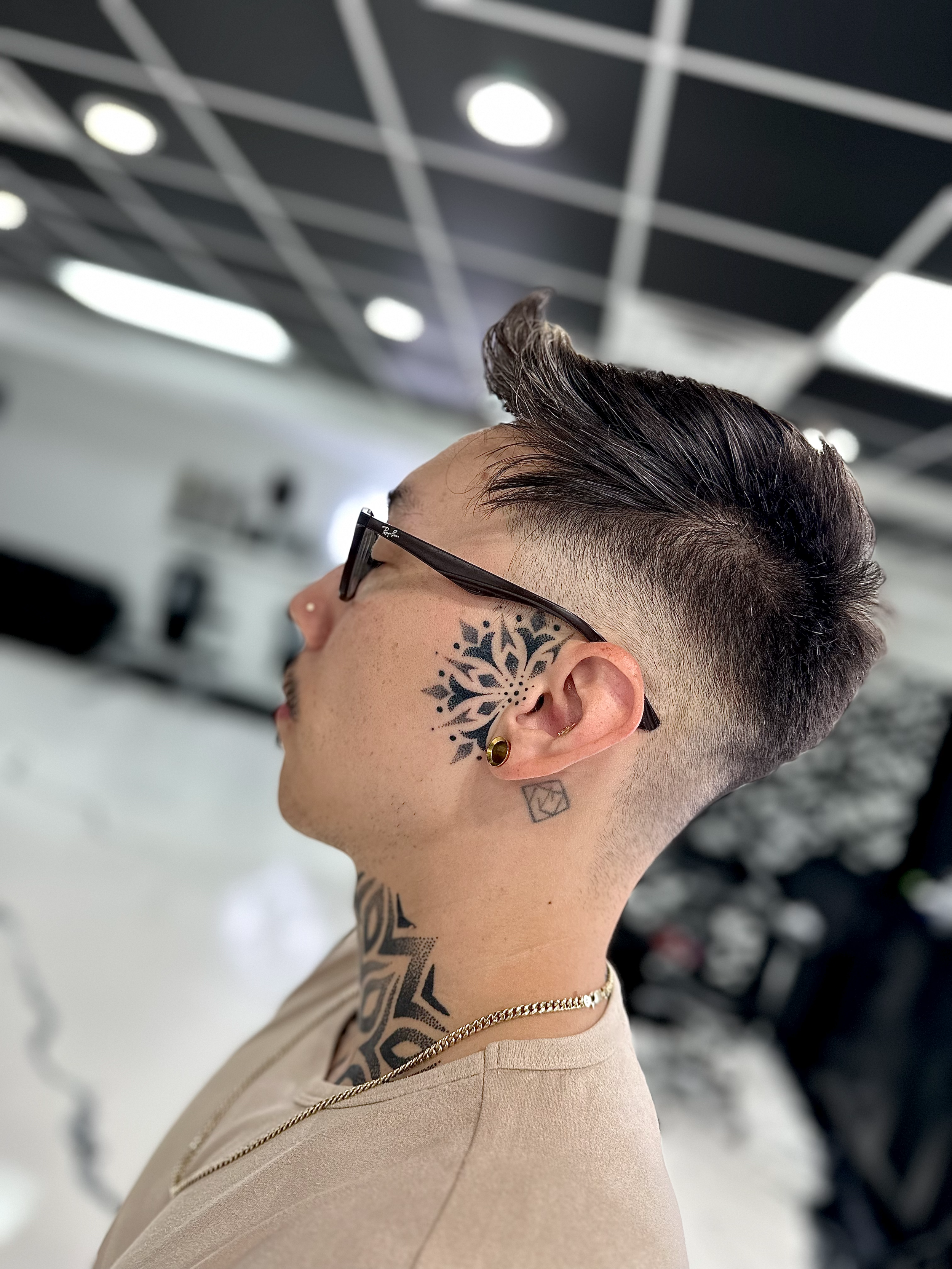 Side view of a person's head showing a hairstyle, tattoos, glasses, and earrings in an indoor setting.