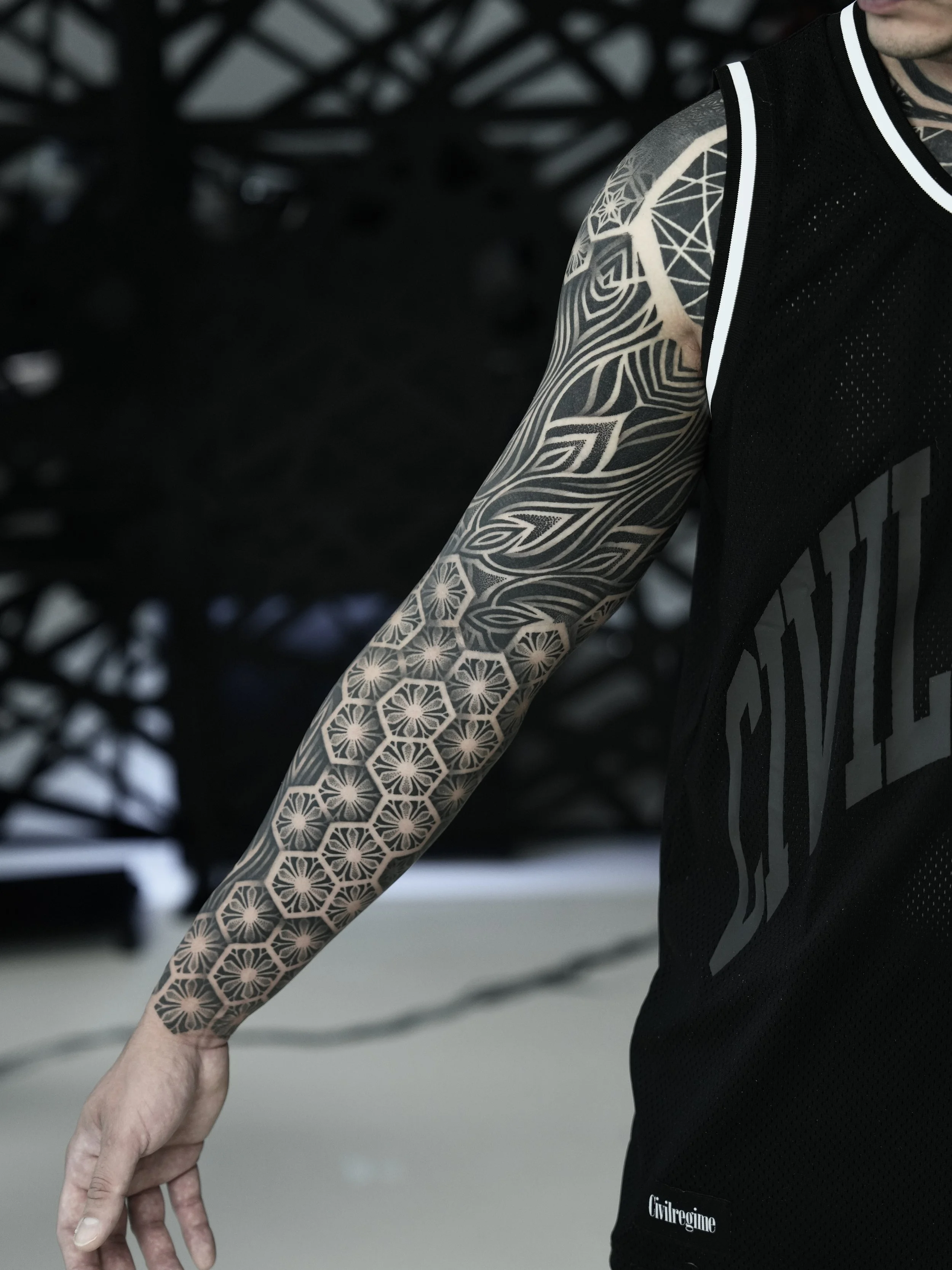 Close-up of a person's arm and shoulder with intricate geometric black and gray tattoo designs, wearing a black sports jersey with white trim.