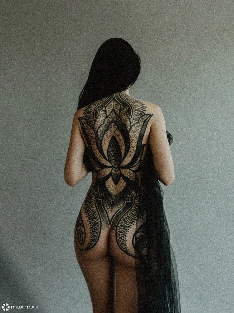 A woman with long dark hair has an intricate black tattoo on her back and lower body, standing against a neutral background with a black fabric draped over her left arm.