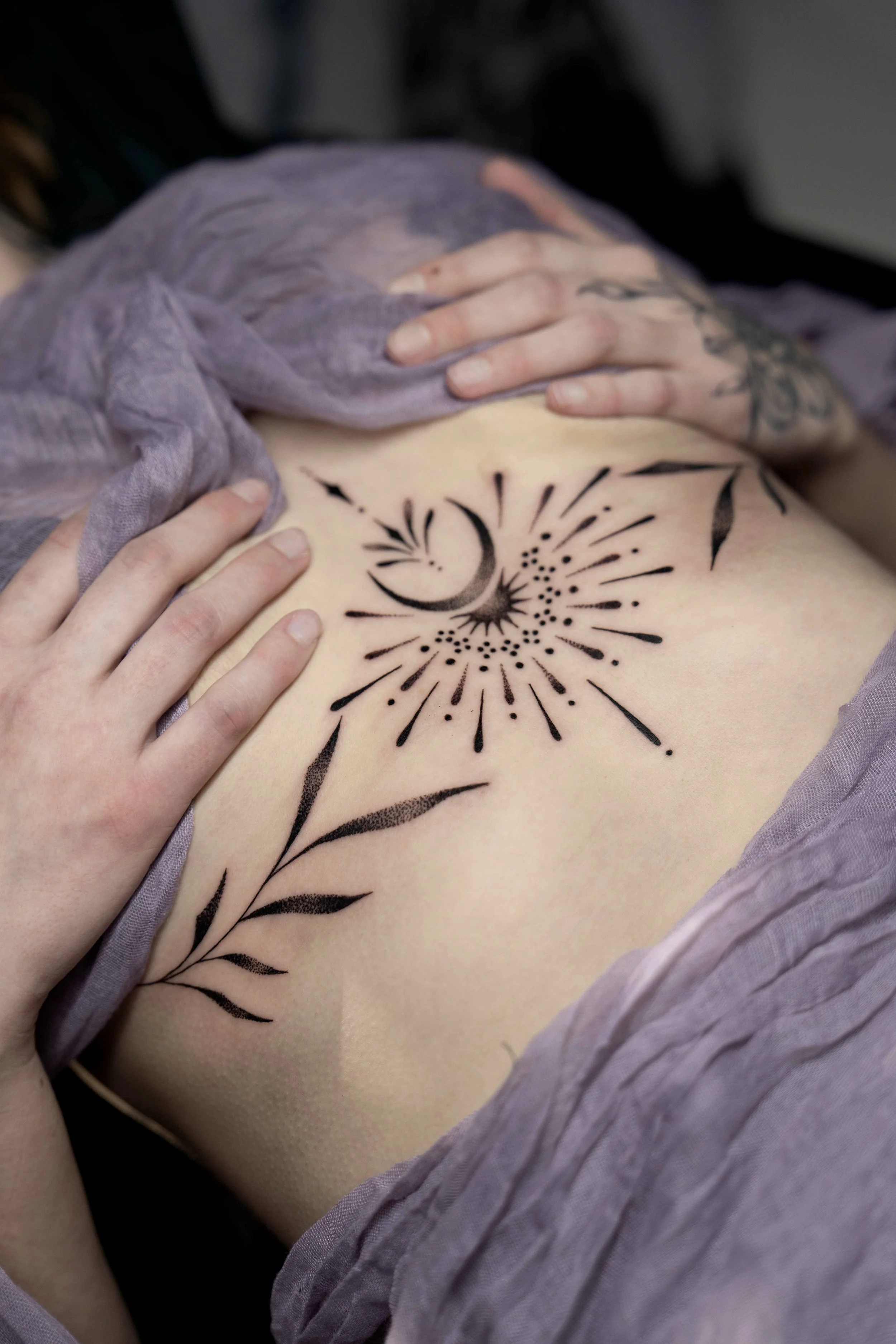 Tattoo of a crescent moon, star, and bursts with lines and dots on a person's midsection, with hands gently touching the tattoo and wearing purple clothing.