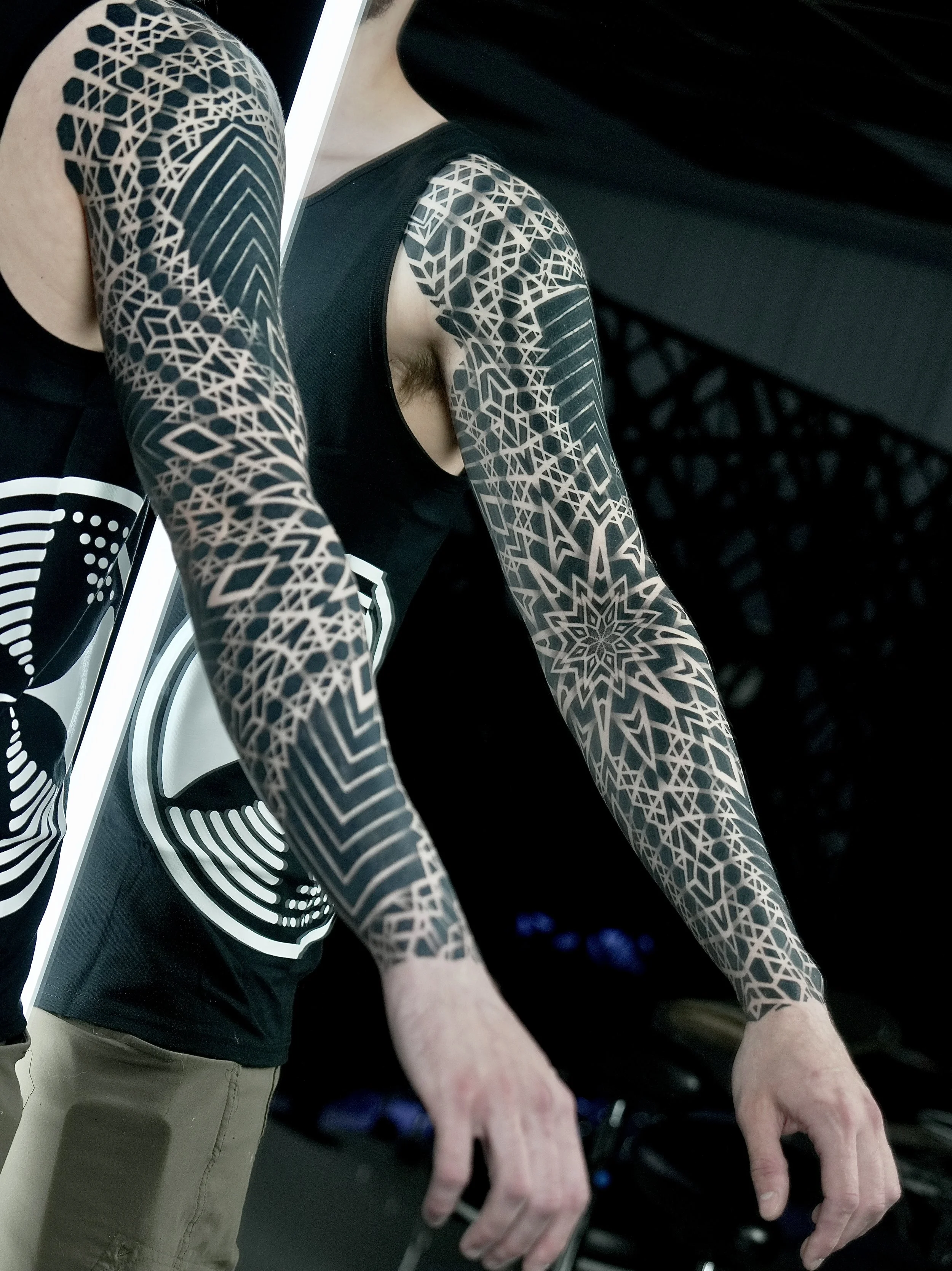 Person with intricate black and white geometric tattoos on both arms, wearing a black tank top with white circular design, standing indoors.