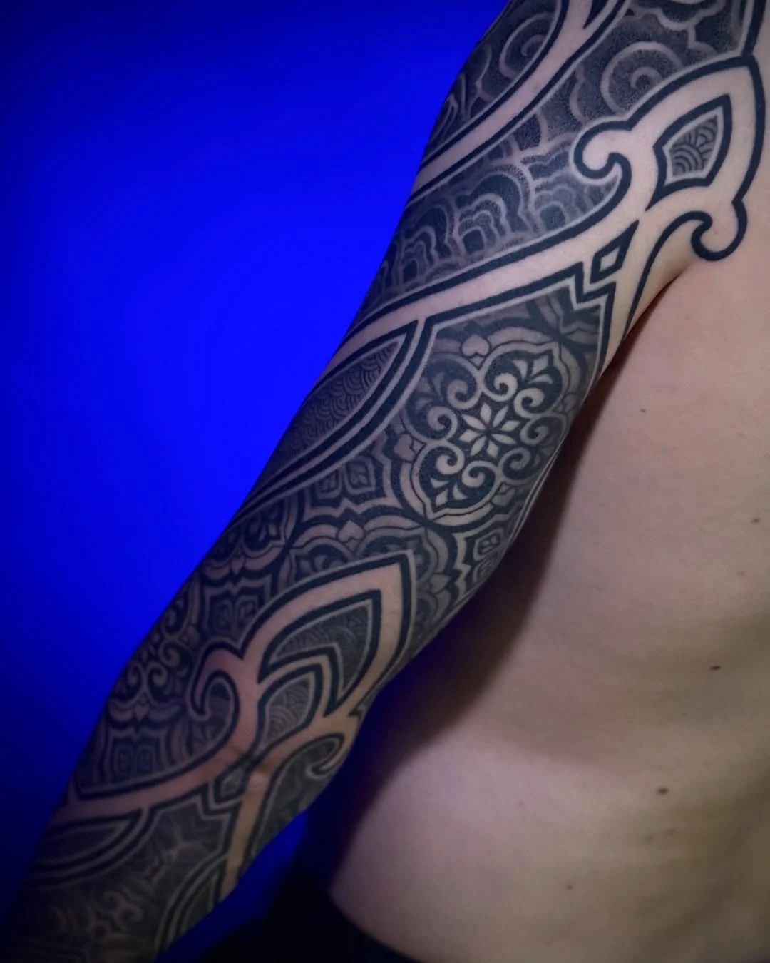 Close-up of a person's arm with an intricate black tattoo design, against a blue background.