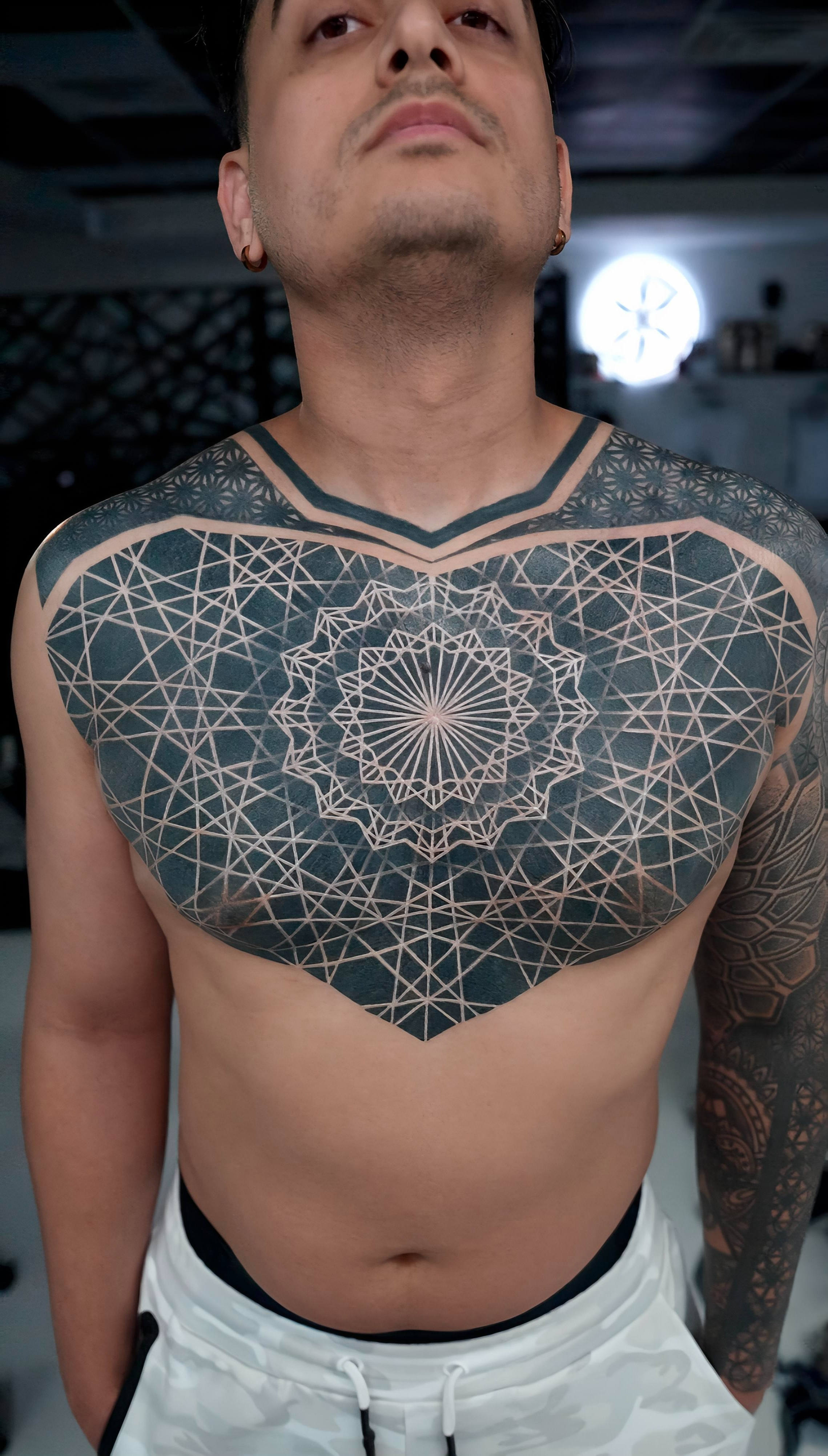 A man with tattoos on his chest and arm poses shirtless indoors.