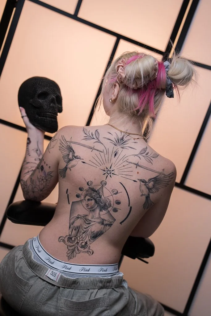 A woman with colorful pink and blonde hair styled in buns with hair ties and a stick pierce, showing her tattoos and holding a black skull decorated with beads or crochet. She is topless, wearing beige pants and white underwear with black text, and i