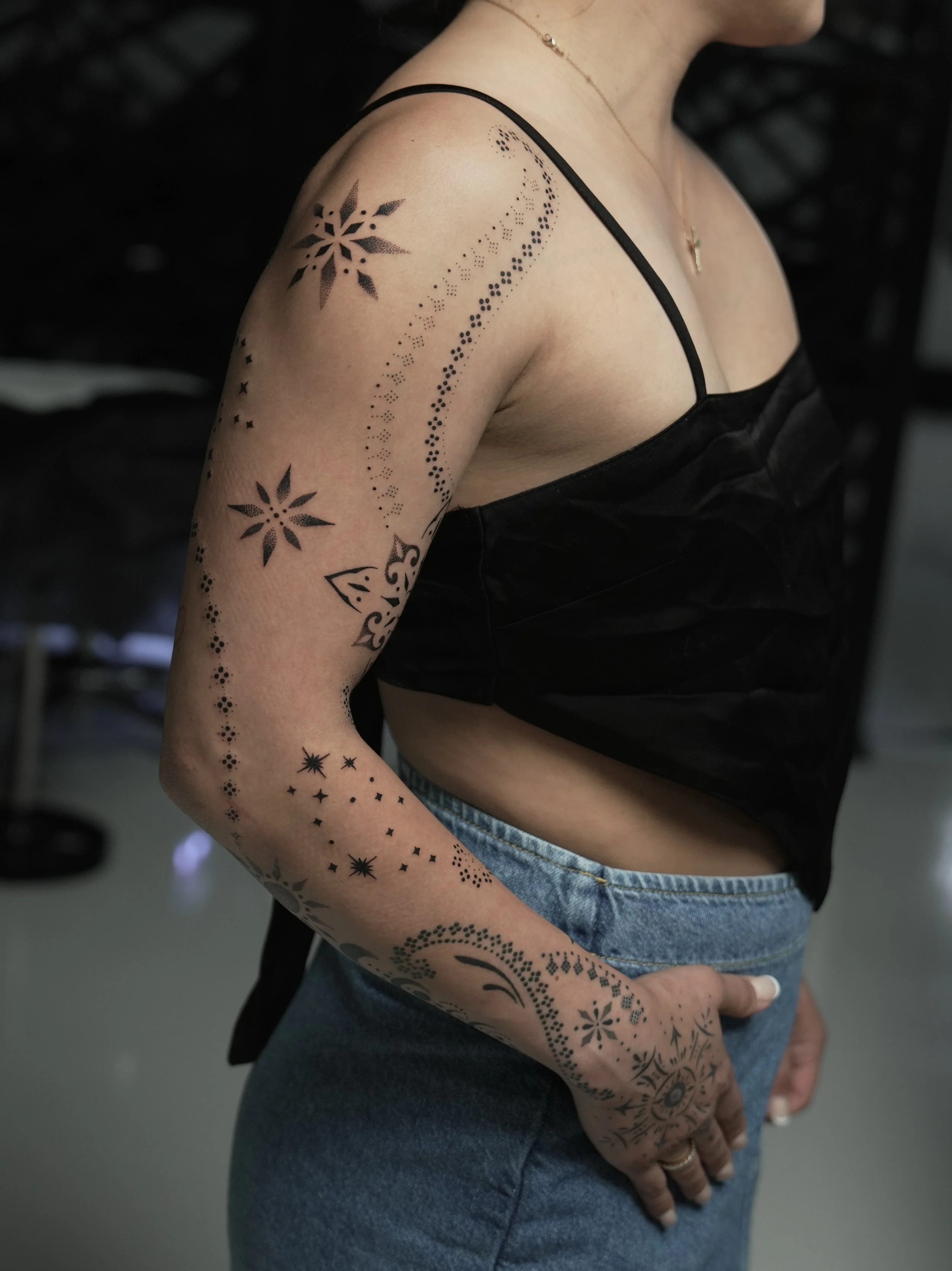 Woman with tattoos on her arm, including floral and celestial designs, wearing a black spaghetti strap top and blue jeans.