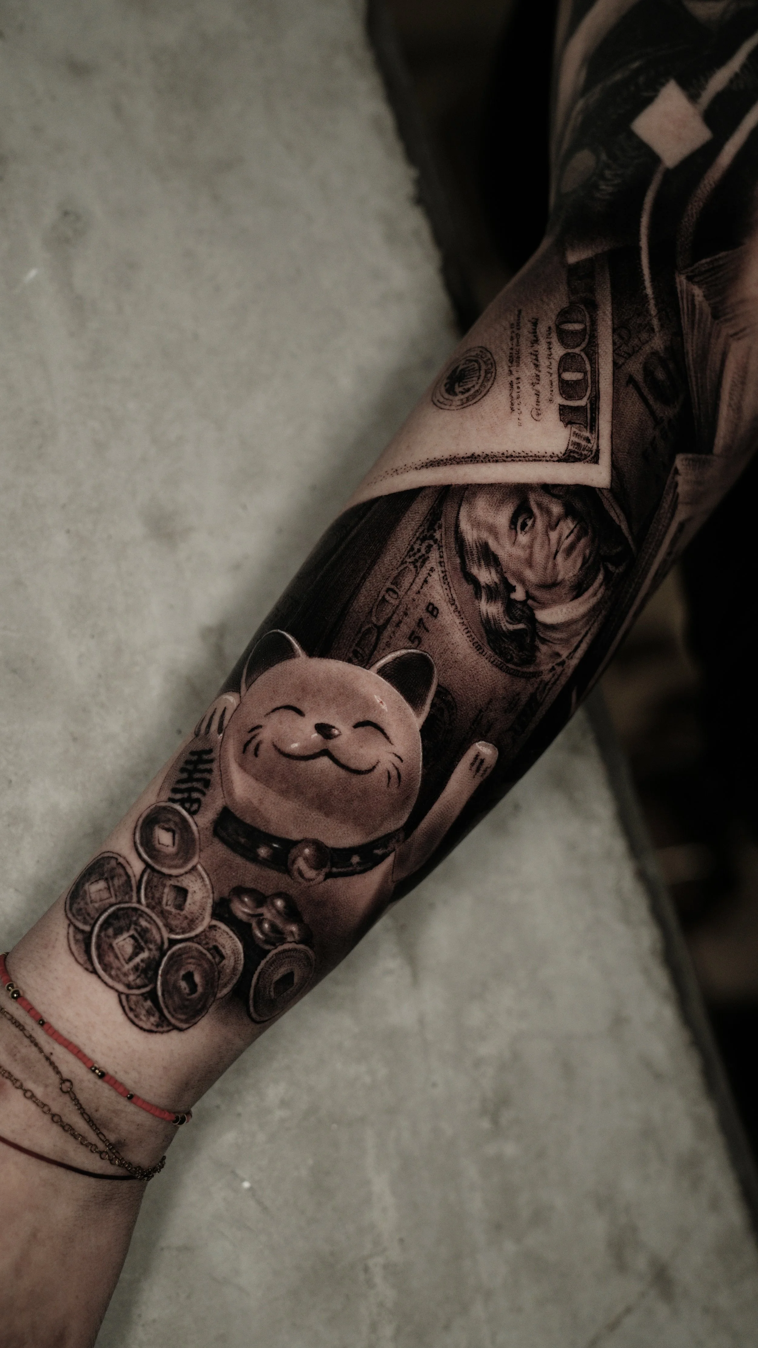 Tattooed arm with a Maneki-neko cat figure, a cartoonish cat with a big smile and a raised paw, along with a tattoo of a U.S. dollar bill featuring Benjamin Franklin.