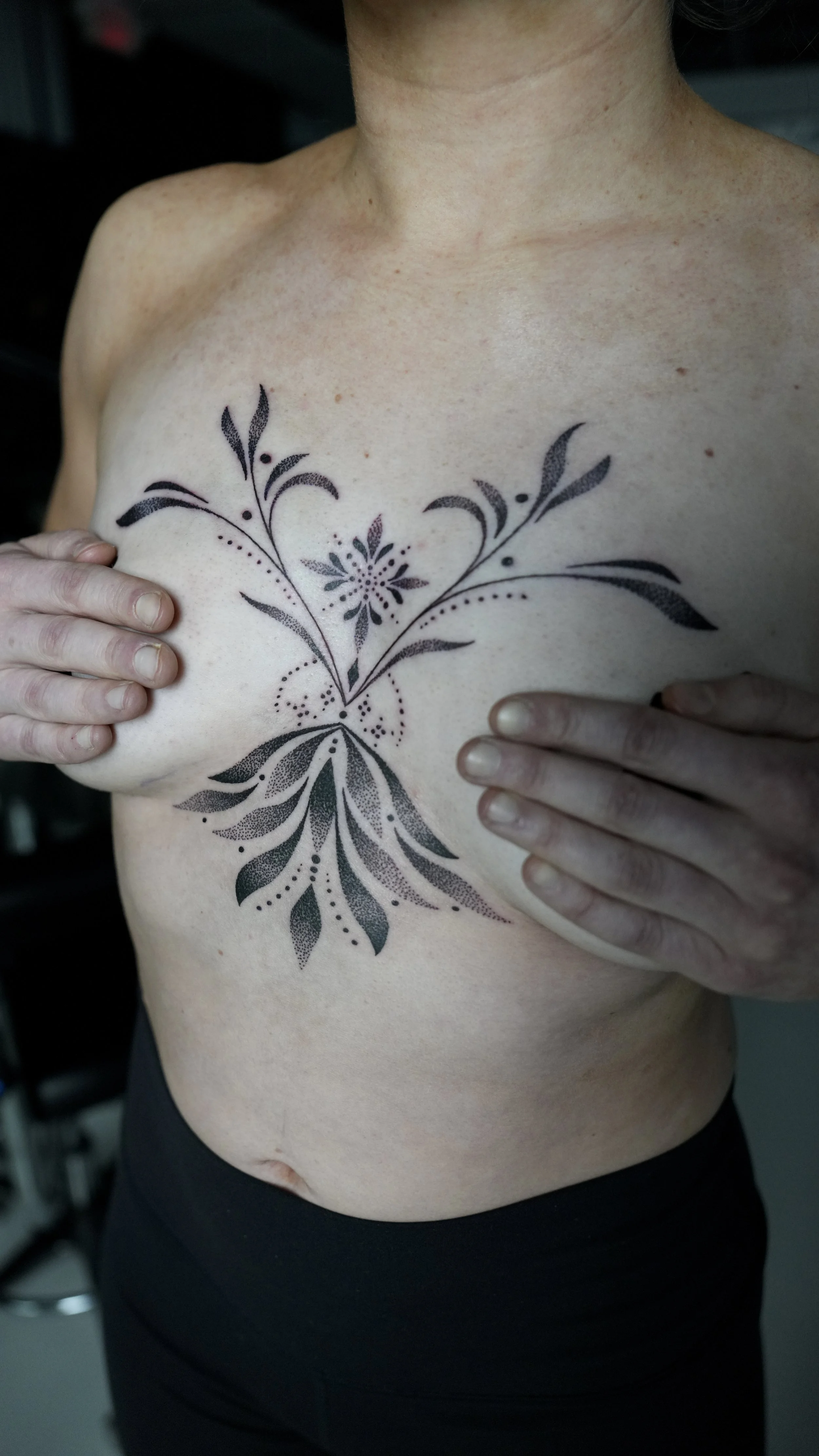 A person with a floral tattoo on their chest, covering part of their breasts, with hands holding their breasts, wearing black pants.