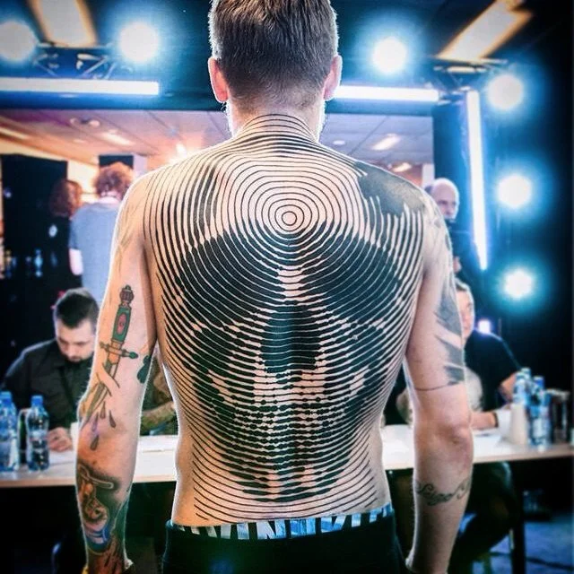 Man with tattooed arms and back, showcasing a large spiral art tattoo forming a skull on his back, in a lively indoor setting with bright lights and people sitting at tables.