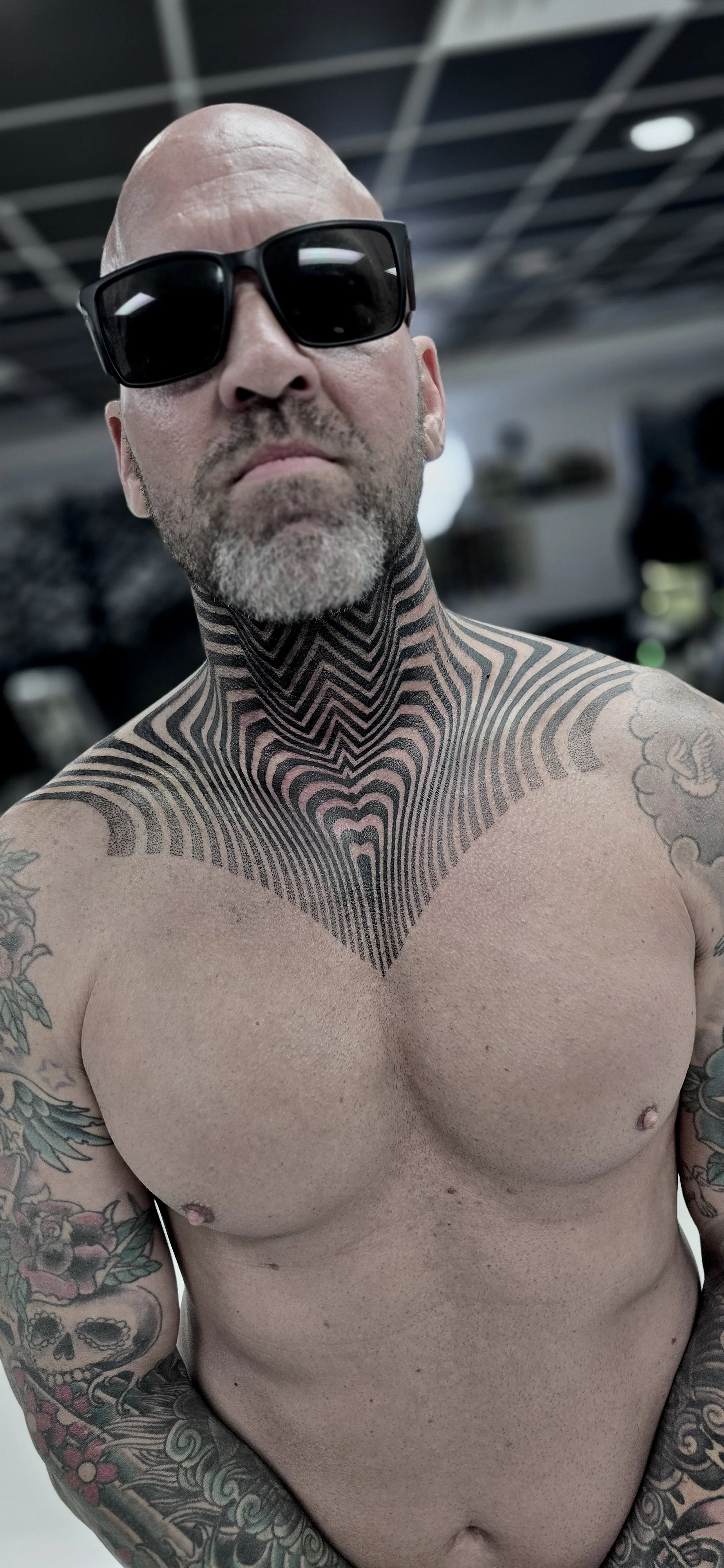 A shirtless man with glasses and a beard taking a selfie, showing tattoos on his chest, neck, and arms, with a blurred indoor background.