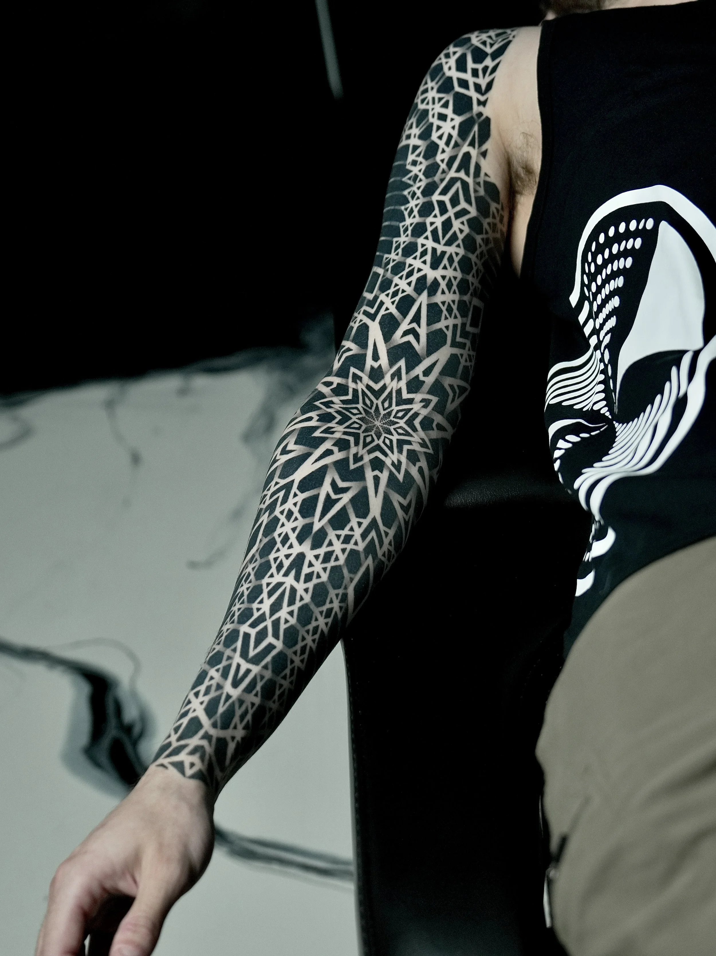 A person's arm with a detailed geometric black and white tattoo, wearing a sleeveless black t-shirt with a white graphic design, sitting in a black chair.