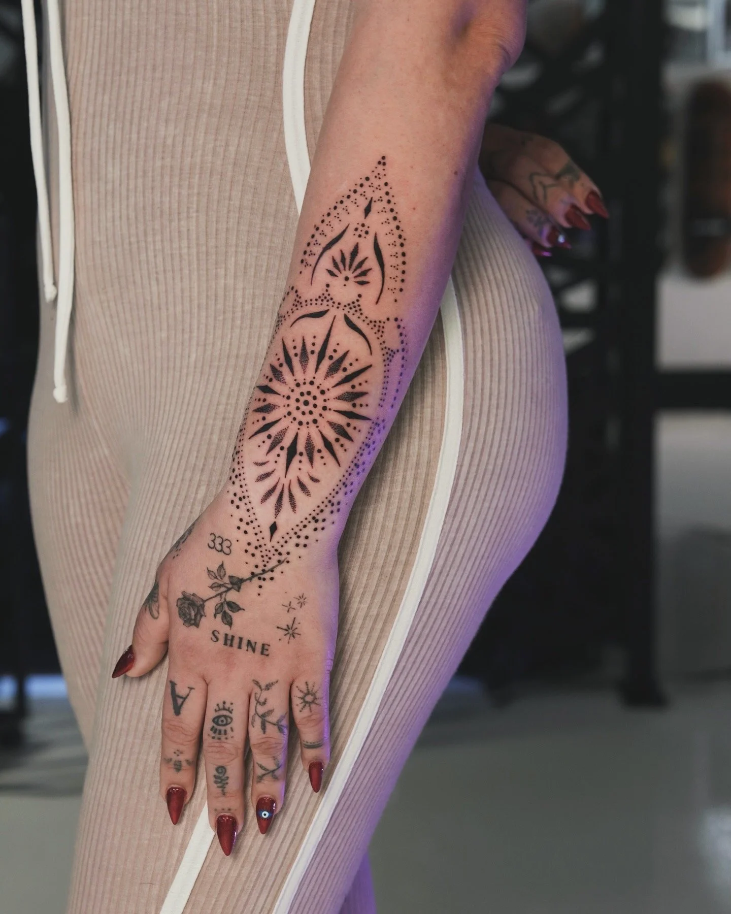A person's right arm and hand with multiple tattoos, including a large geometric pattern on the forearm, a rose design on the hand, and various smaller symbols and words on the fingers and wrist, with beige clothing.