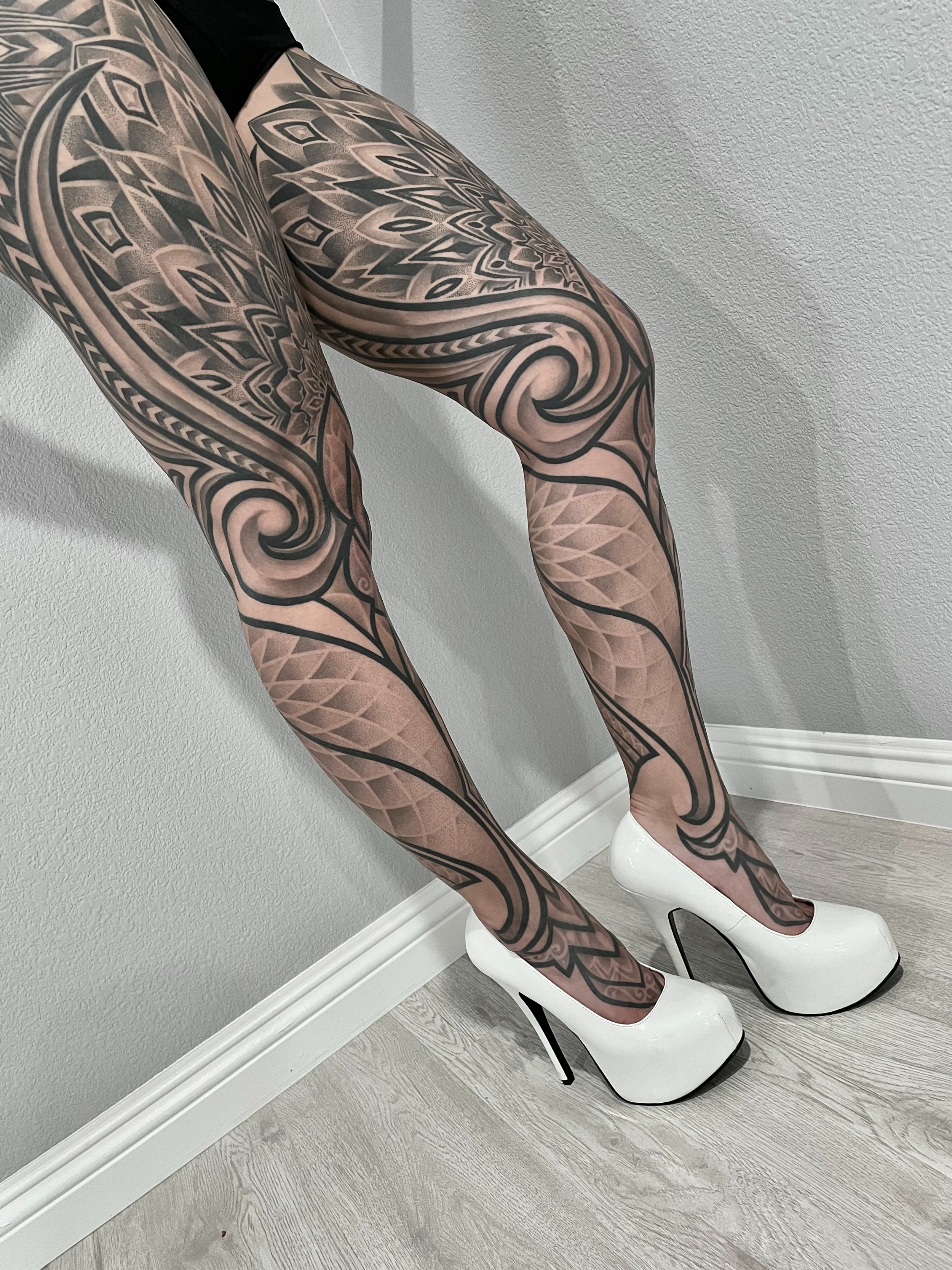 Close-up of a person's legs with intricate black and gray patterned tattoos, wearing white high-heeled shoes, standing against a gray wall and light-colored wooden floor.