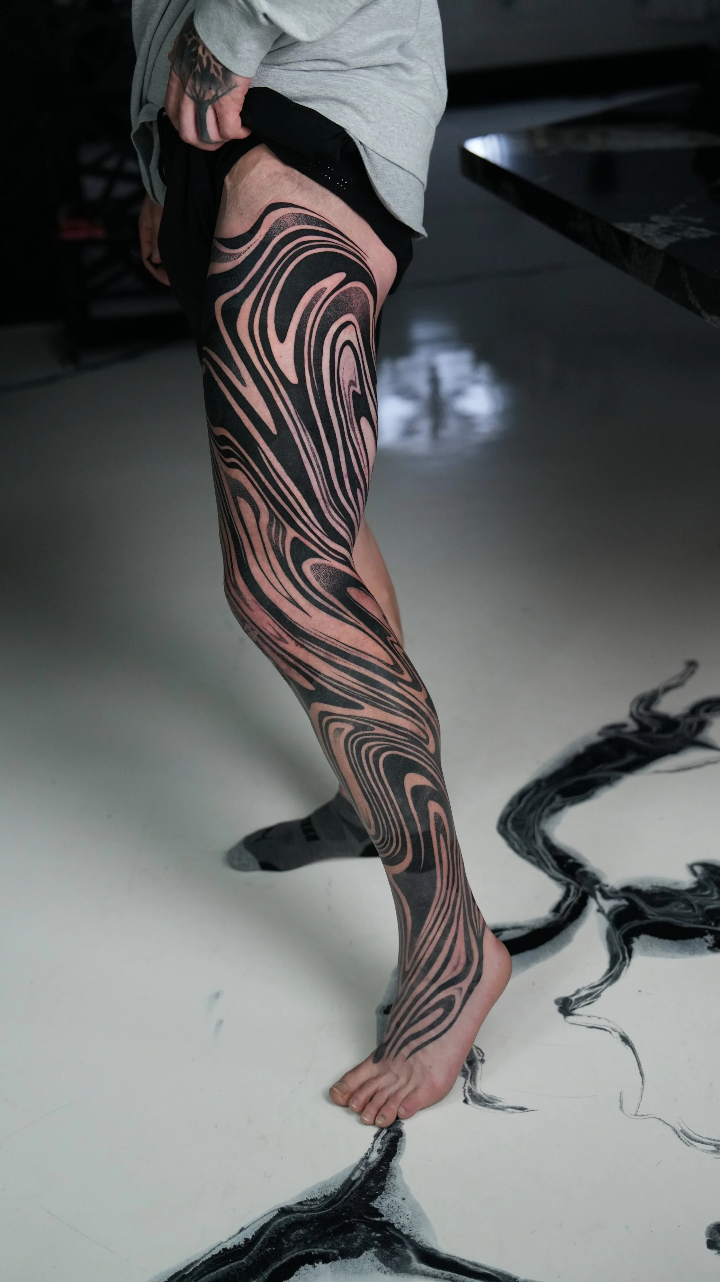 A person's leg with a large, black and pink swirling tattoo extending from the thigh to the ankle, standing on a floor with black and white abstract patterns.