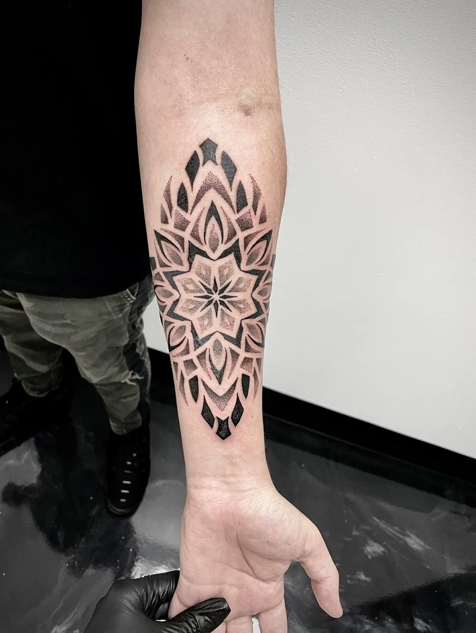 A person's forearm with a large black geometric tattoo featuring a symmetrical pattern with a central star-like shape surrounded by leaf-like designs.