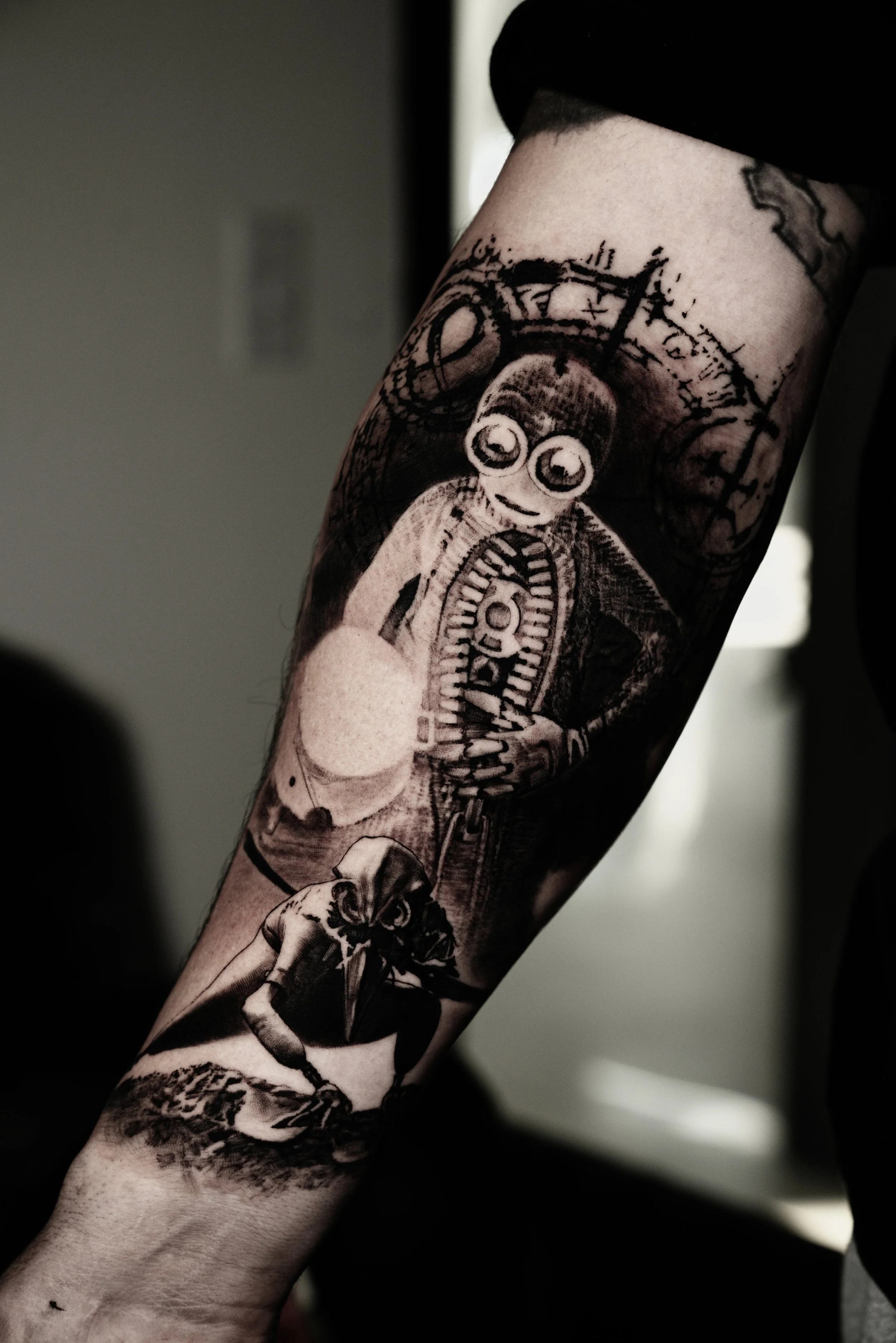 Close-up of a person's arm with several tattoos, including a cartoonish face with big eyes and a skeleton figure, in black and gray ink.