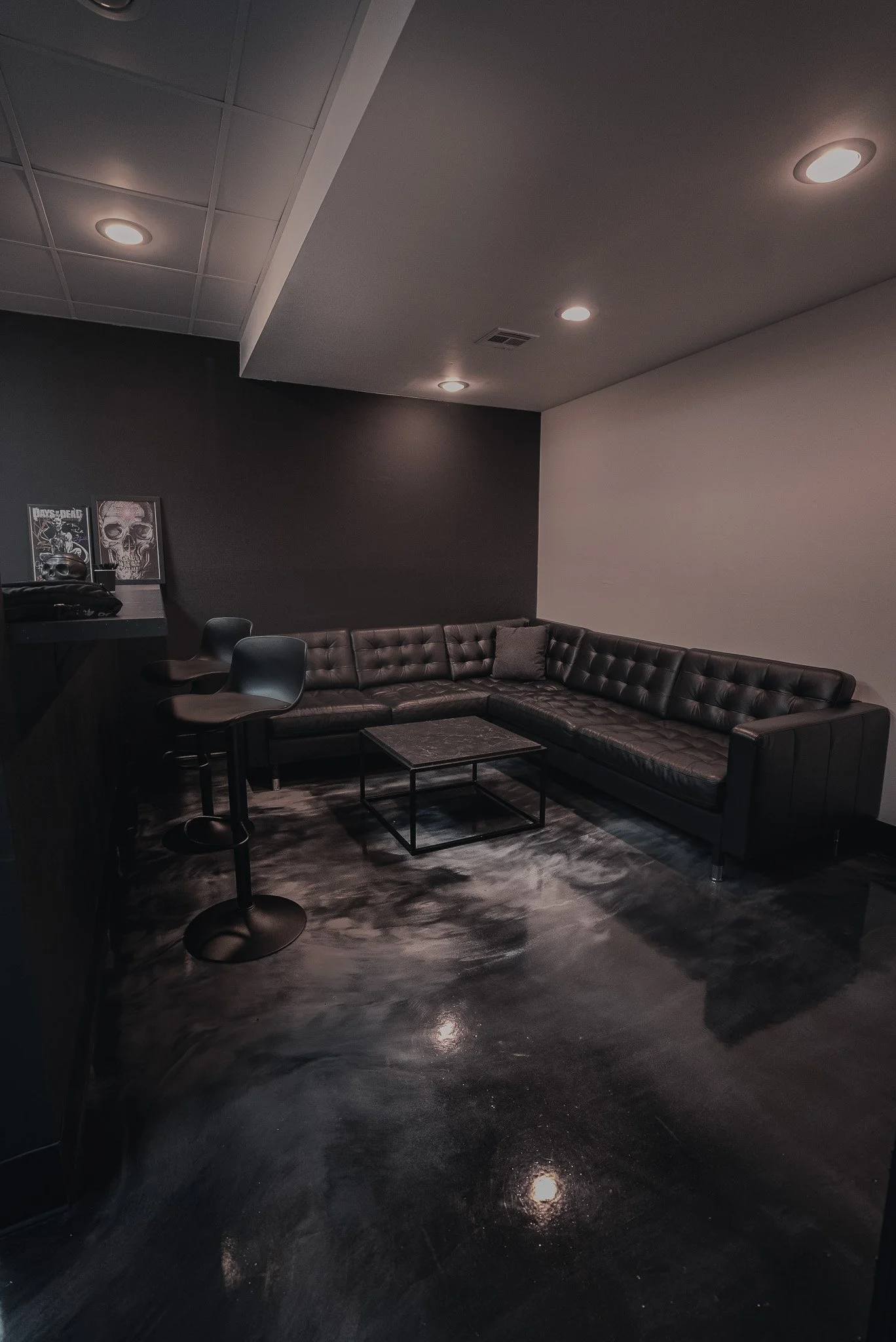 A modern, dimly lit lounge with a black leather sectional sofa, a small black coffee table, two black bar stools, and dark walls adorned with artwork, all on a polished dark floor.