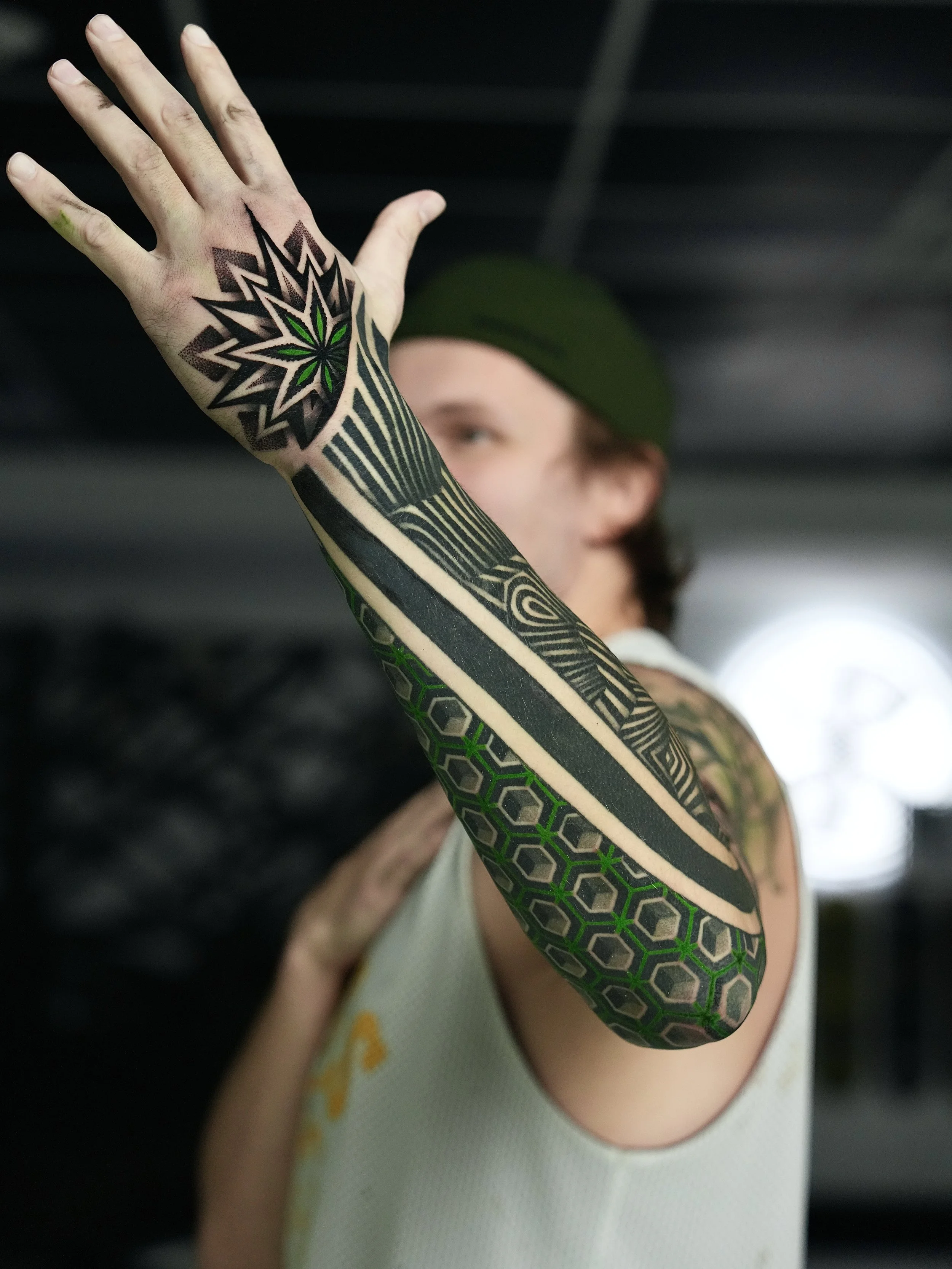 Person with intricate black and green geometric tattoos on his arm, wearing a sleeveless athletic shirt and a green cap, extending his arm forward indoors.