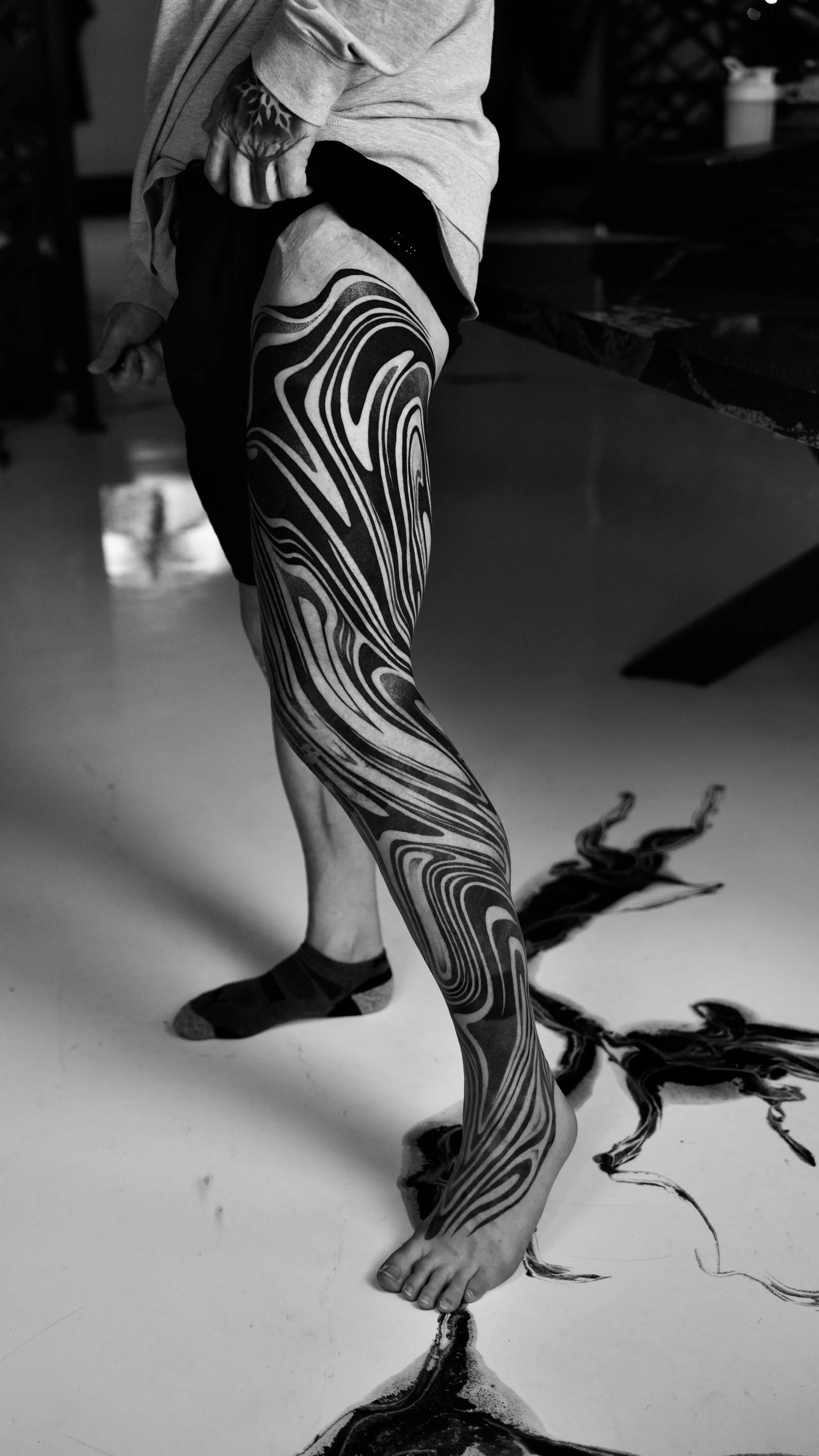 Person with tattooed leg standing on a floor, with black ink spilled on the floor creating a phoenix design.